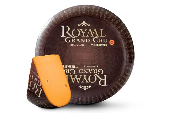 A large round wheel of cheese wrapped in dark brown packaging, with a wedge of orange cheese cut out and leaning in front. The wrapper has large gold lettering that reads “ROYAL GRAND CRU” and smaller text “BY BEEMSTER.” A small red-and-gold circular seal appears to the right of the word “CRU.” The wrapper also has a faint illustration of a cow in the center and repeating small “ROYAL” text around the outer edge.