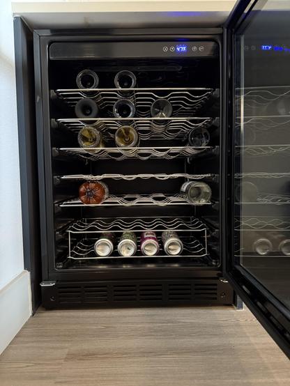 Wine fridge with door open exposing random bottles with some cans of sour smoothies on the bottom