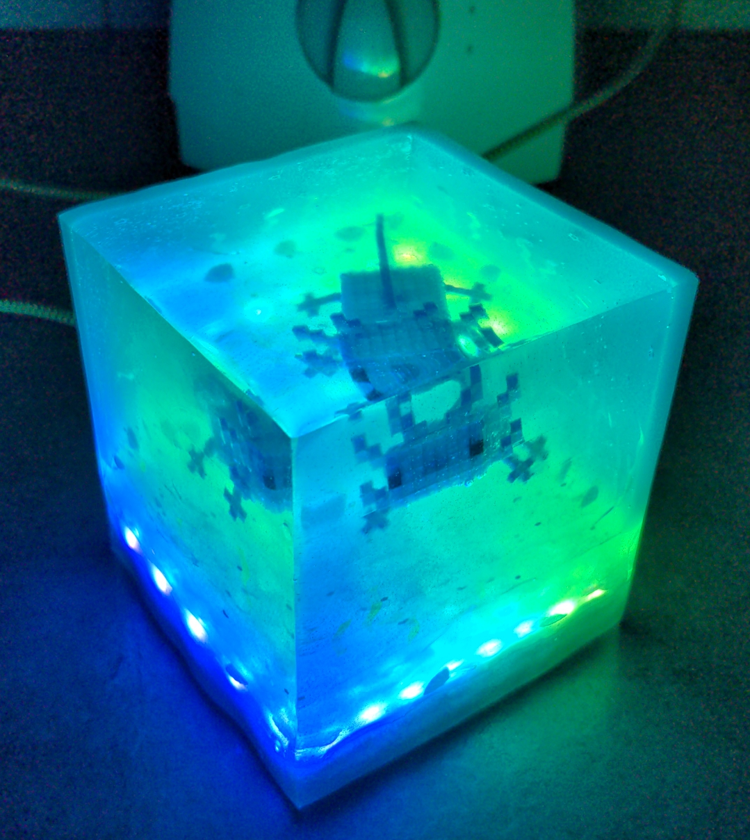 a green and blue glowing epoxy resin cube in the dark with a Minecraft axolotl inside