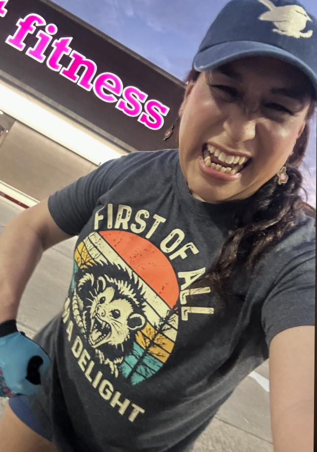 Me, a trans femme Latine, giving you a little taste of the gun show in front of a gym.  Part of the sign can be seen in the background that says "fitness"

I'm wearing a graphic tea that has a possum with its mouth open looking like it's snarling. The words, "first of all I'm a delight on top and bottom of the possum.