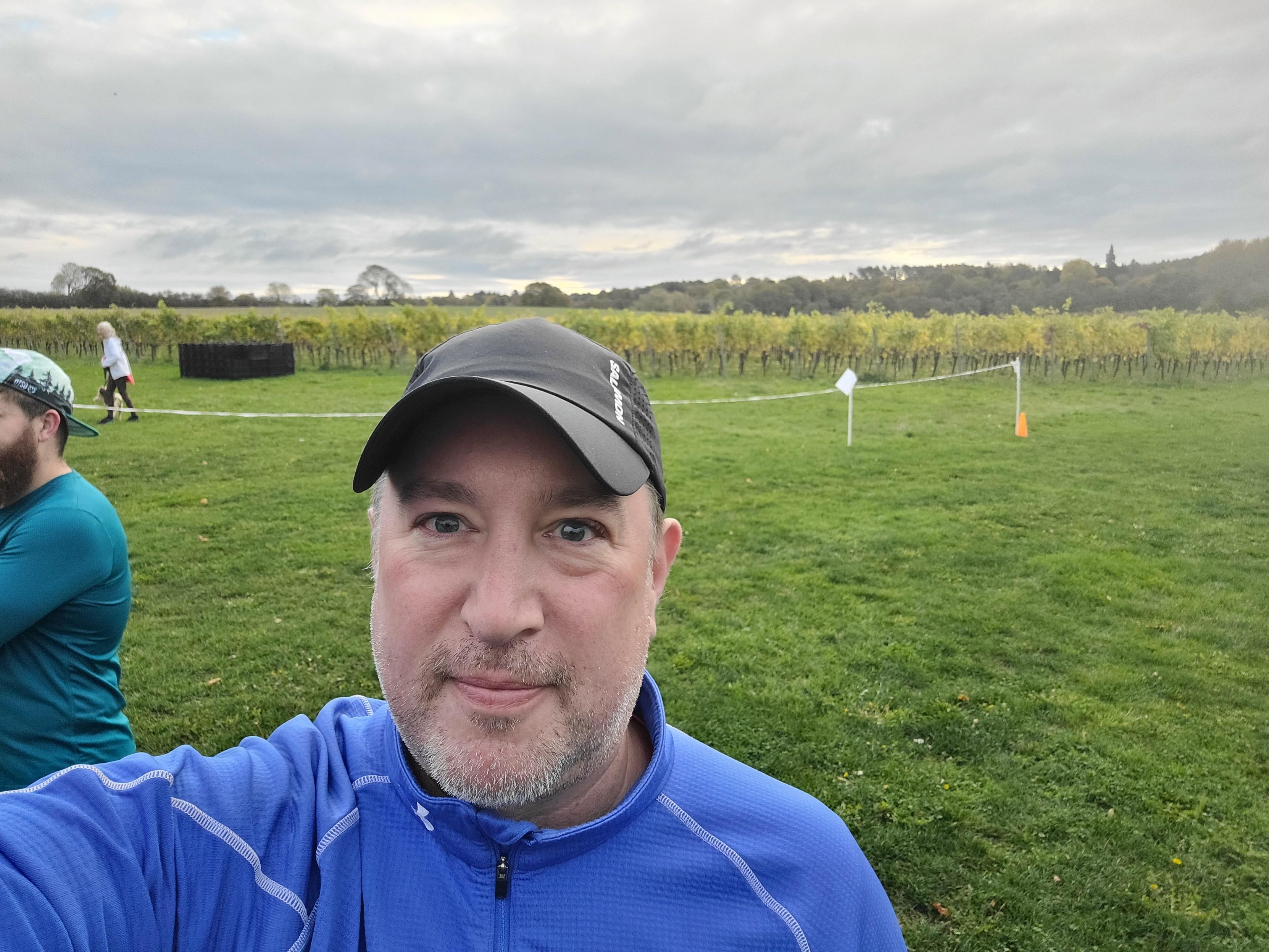 Selfie of my big dumb face with the vineyard in the background and runners milling about. I've got a blue sports jumper on and a tiny hat that makes my head look like a thumb. I've got some beard scruff going on that appears to have grown back white for some reason probably due to WITCHCRAFT because I'm a young stallion in the prime of his life (in my mind). I look like a kindly old gnome king, banished from his land. Like a sentient nose that someone has badly crafted a face around in plasticine. Like a child who's been zapped with a raid-ageing (aging?) ray and doesn't understand what's going on. Or perhaps a dog who's been transmogrified into human form. The sort of person who you might ask directions from, but they'd send you the wrong way by mistake. I've got the sort of face you might expect to see if you're on an adventure and need clues from a friendly wizard found on the path. I look, frankly, fucked. 