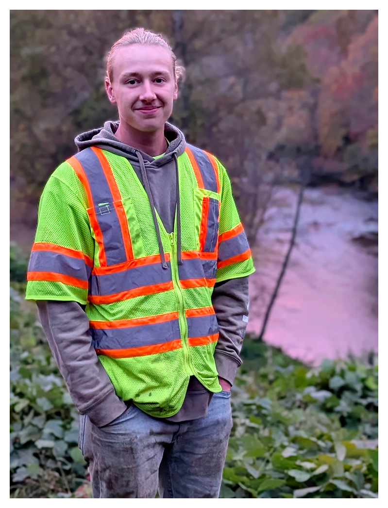 a young man with blond hair tied back, wearing a bright yellow safety vest with orange and silver stripes over a gray hoodie, stands outdoors in green foliage and a river with trees in the background, hands in pockets, smiling slightly.