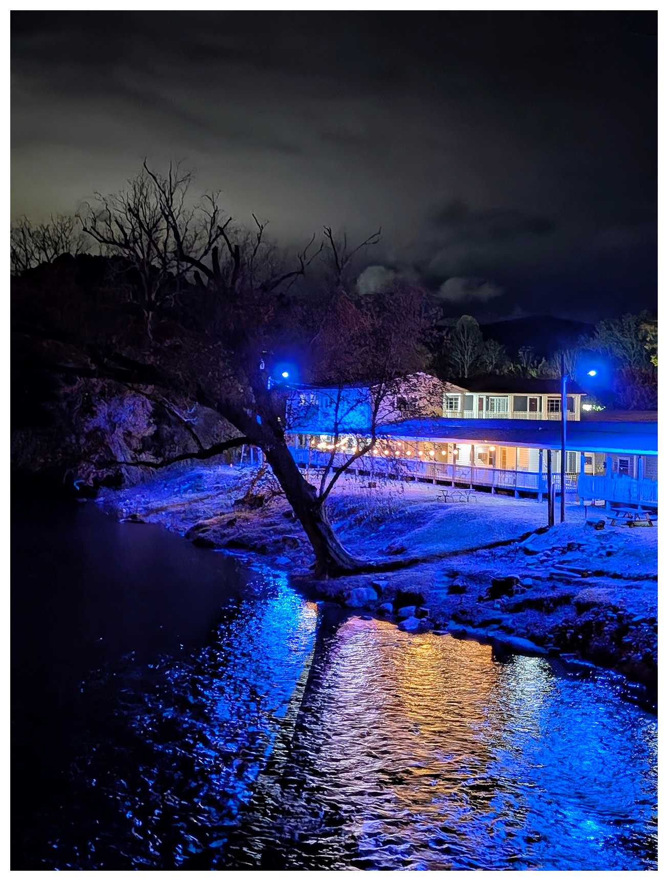 ai-assisted alt text:
	
A warmly lit building sits by a river at night, surrounded by trees under a cloudy sky. Blue lights illuminate the water’s edge and trees, reflecting brightly on the rippling river. The scene feels quiet and serene with dramatic lighting contrasts.