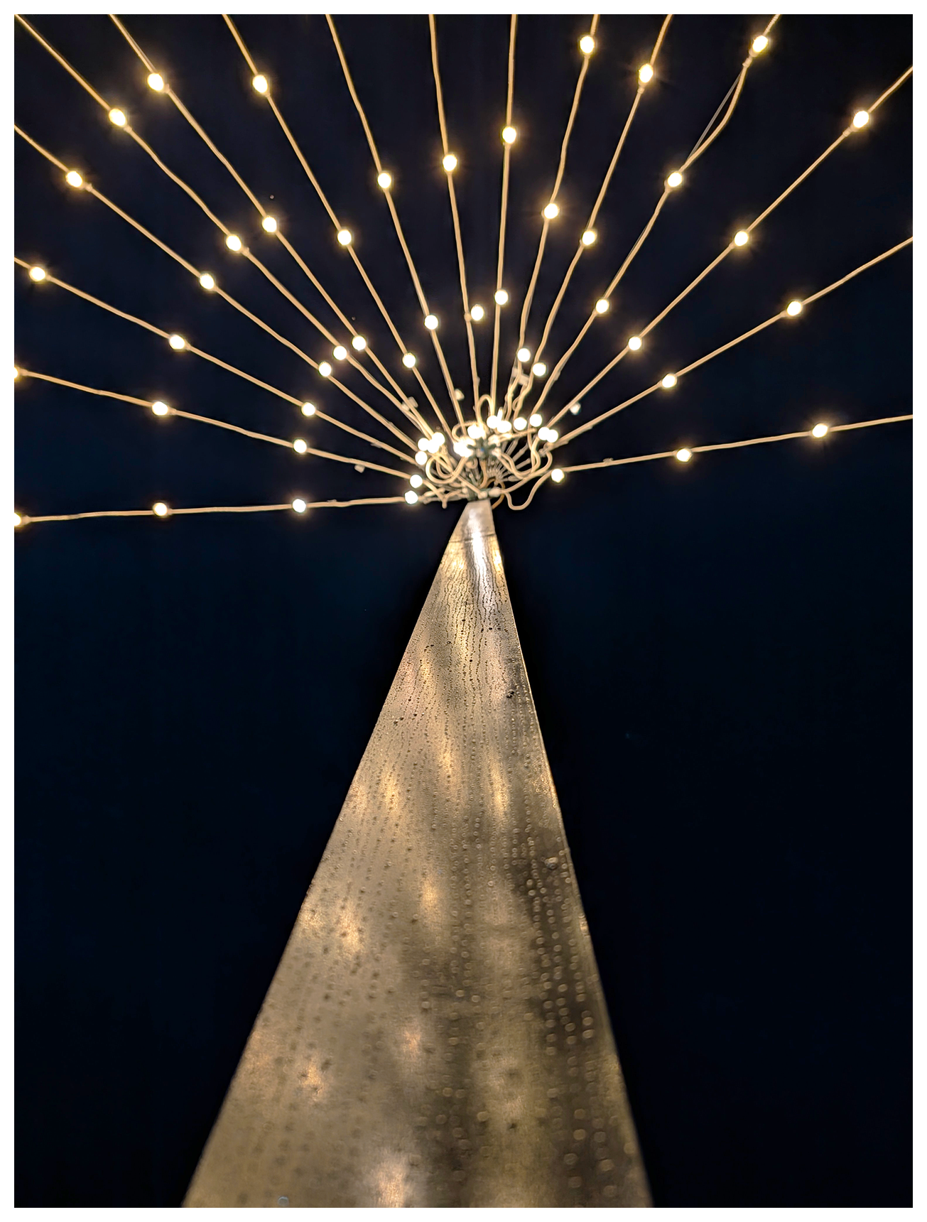 Abstract. view up a rain-spattered metal pole to a cluster of string lights fanning outward in a starburst pattern against a black, night sky.