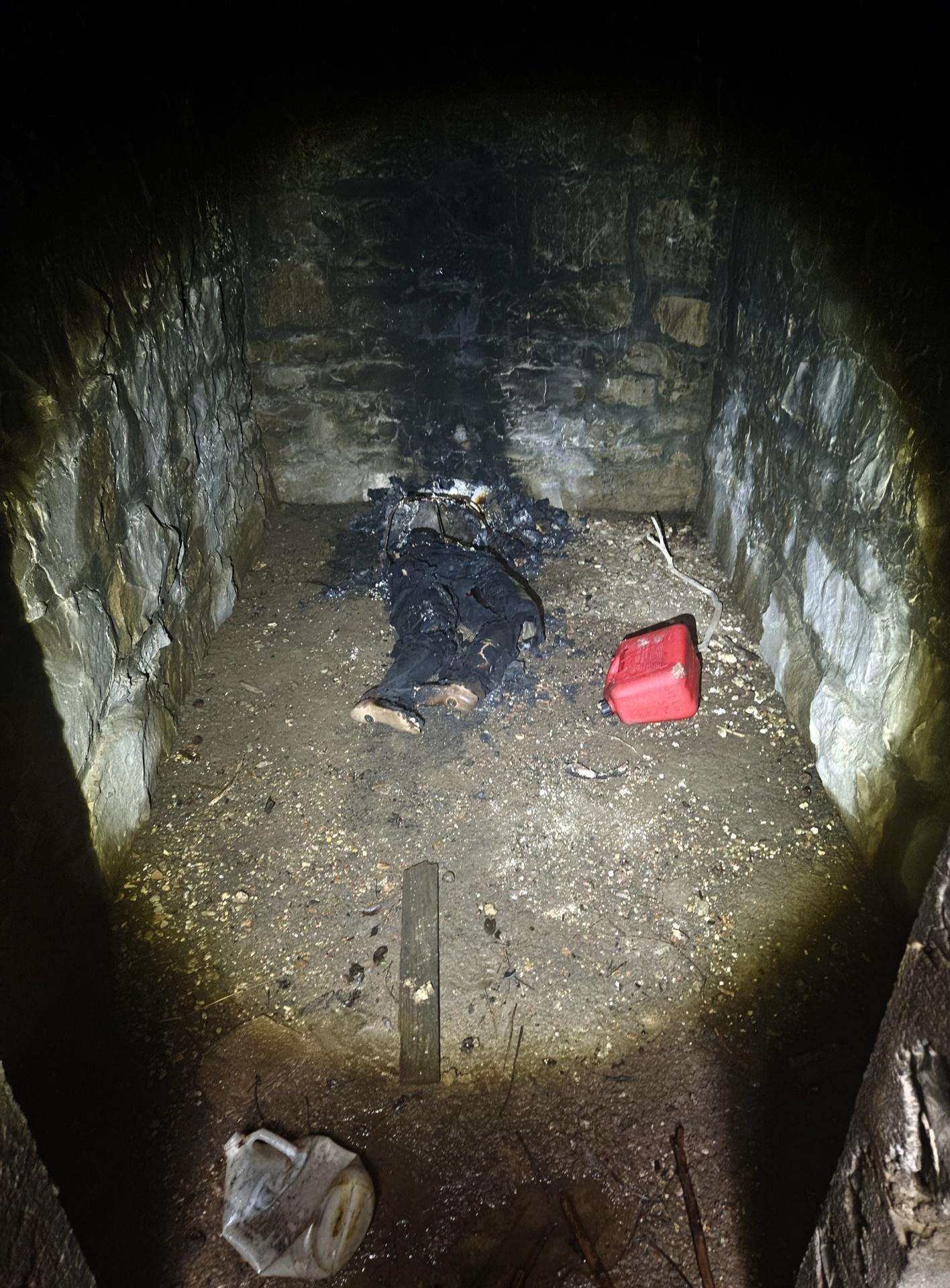 Stone walls with a flashlight beam. You see a red gas cans in the corner, and what looks like a burnt body