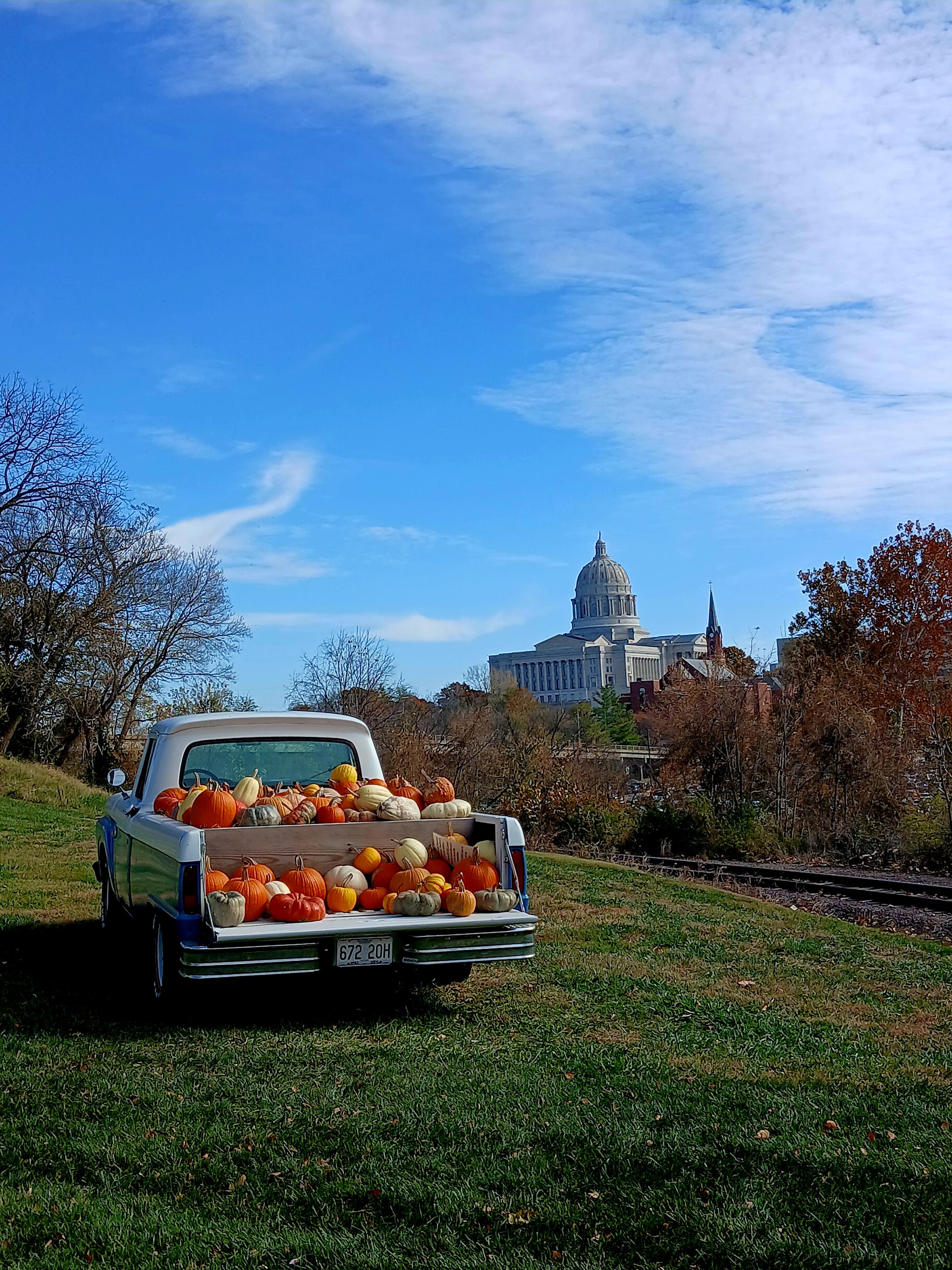 An older truck in blue and white sits next to the train tracks. The bed is filled with Pumpkins and Squashes in orange, white, blue, and yellow. In the background you can see the Missouri state Capitol building