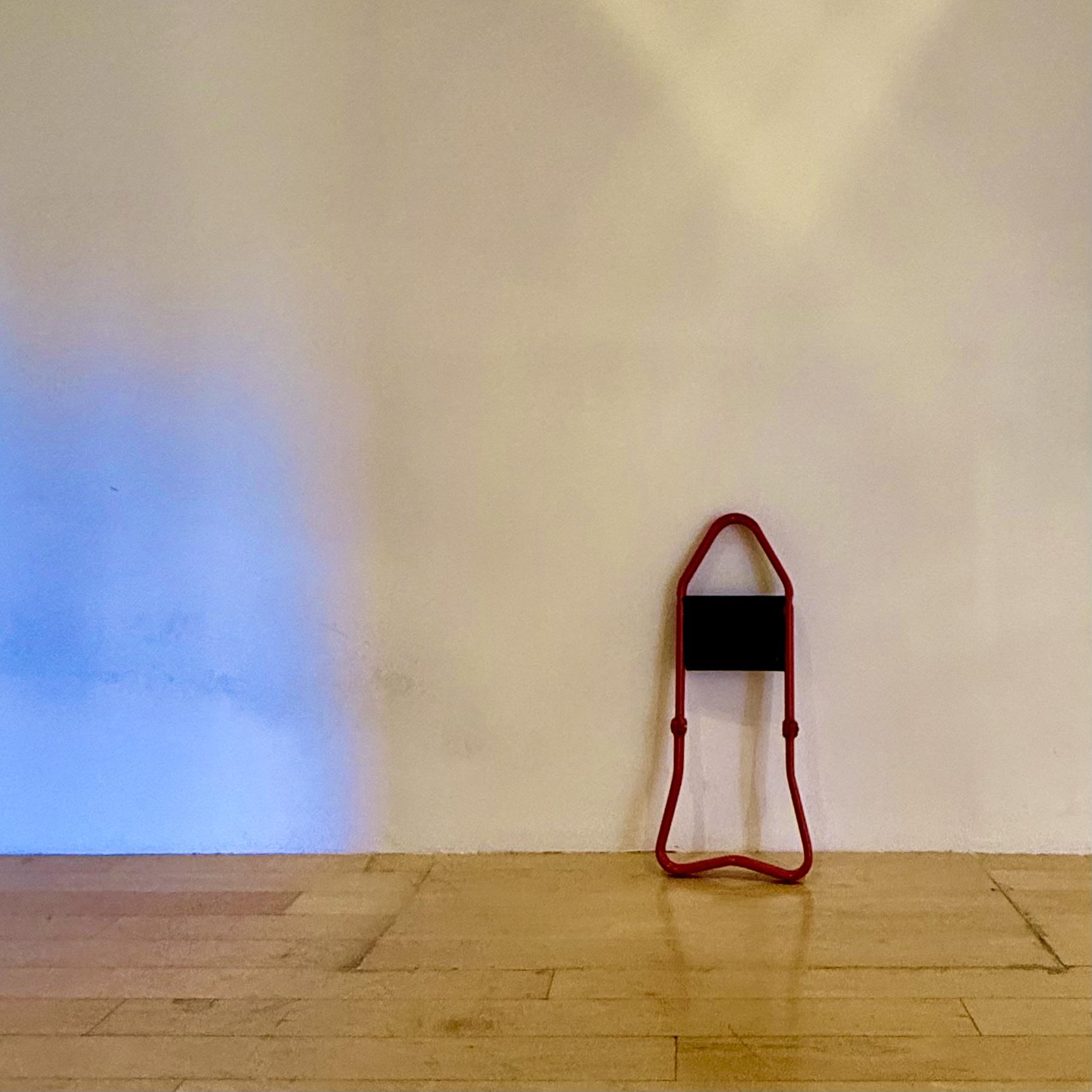 I initially thought this was a step stool, but now I have no idea what it is. It's like a red metal thing that has a hinge and a shelf? Anyway, it's propped against a white wall in an art museum.