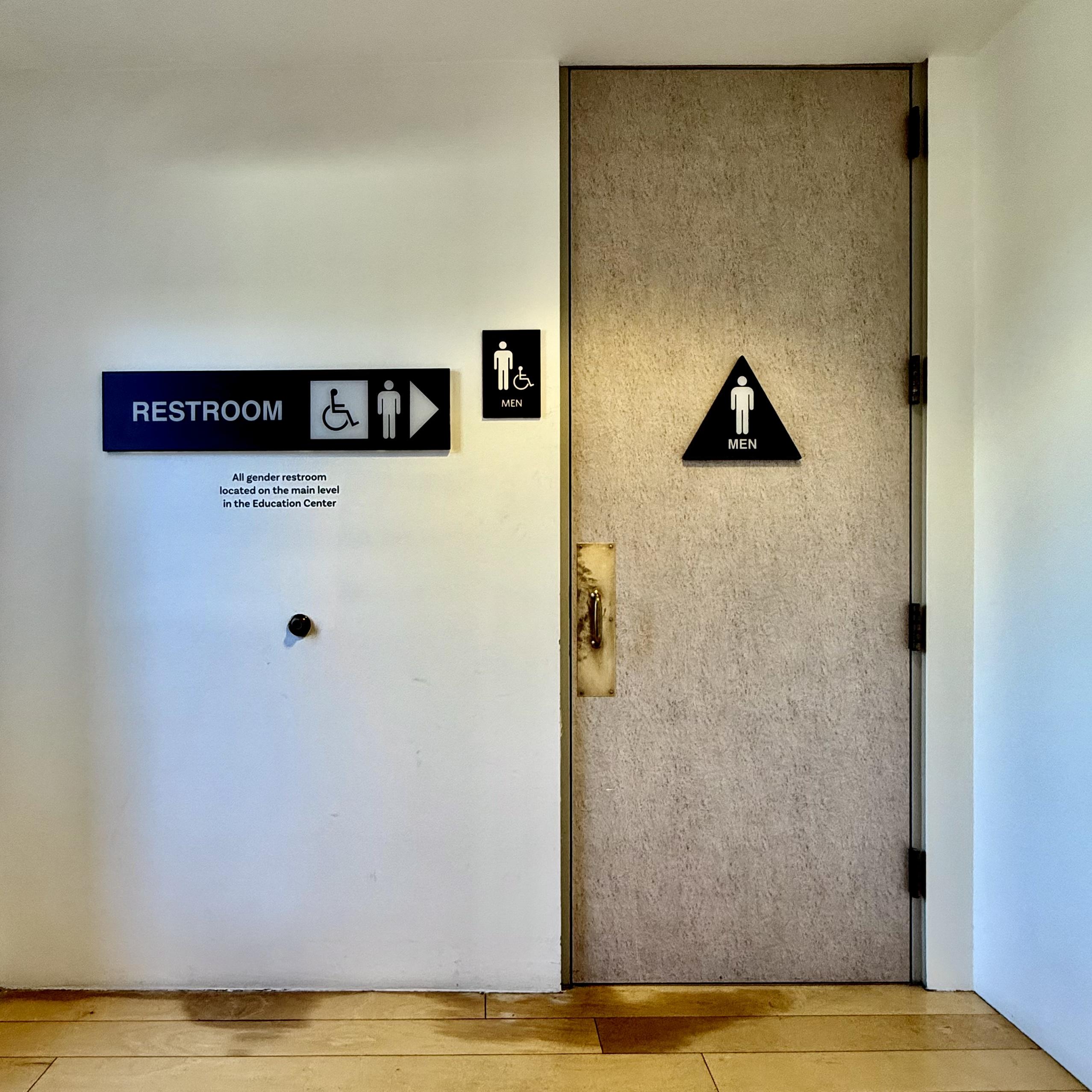 Restroom entrance in an art museum. There appears to have been some flooding in the past because the light wood floors have some water damage and staining.