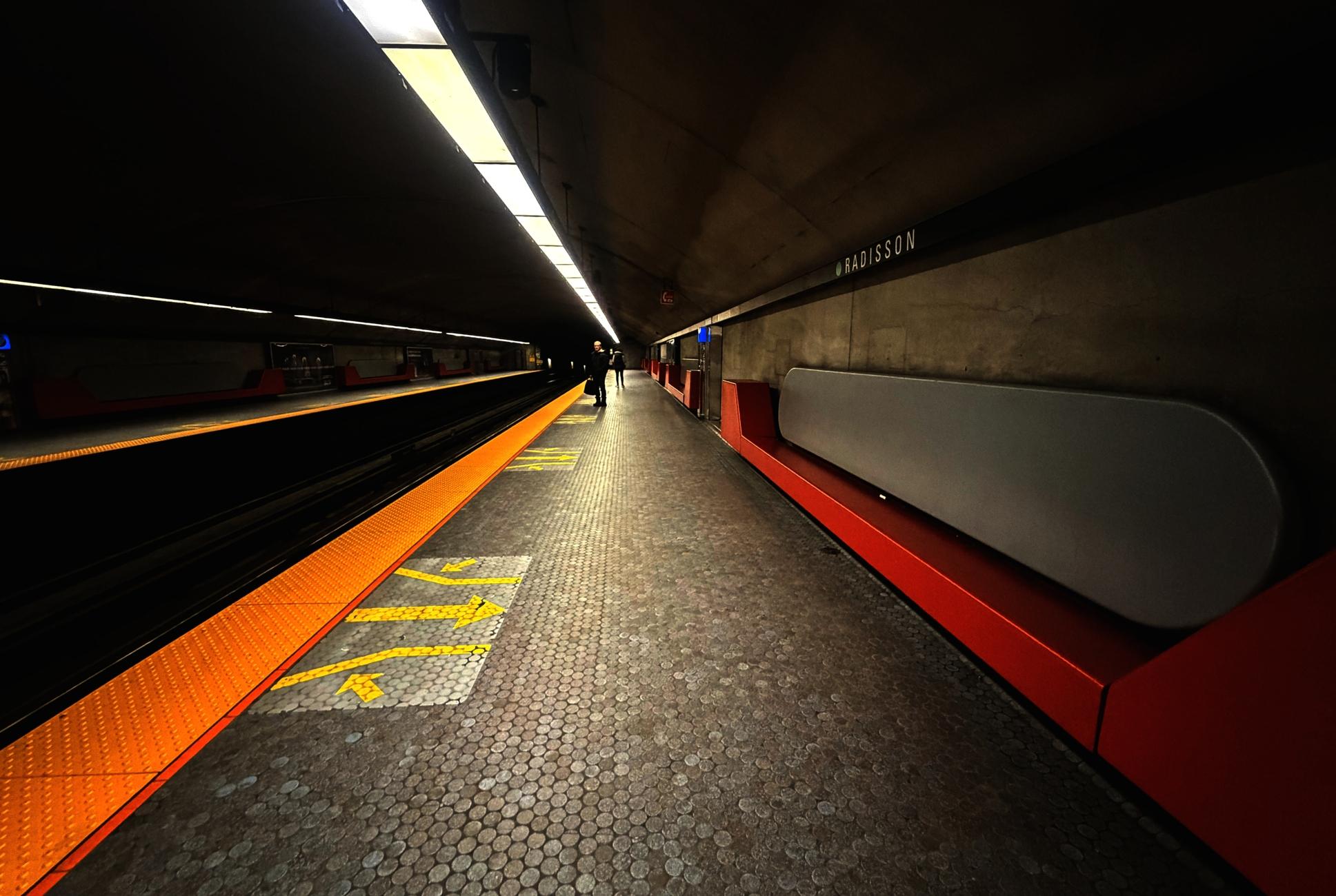 Photograph of an underground metro station, taken from one end of the platform looking towards the tunnel in the back, acting as a vanishing point. On the left, the tracks in the floor, the lights above and the reflective orange surfaces on the platform edges create lines extending into the background. In the center, the platform itself with its small, round blue tiles and yellow arrows indicating the train exit doors, and two passengers waiting at the far end. On the right, along the gray concrete wall, modern-style bench seating, with red seats and rounded gray backs, and the black band at the top with the station name "Radisson" in white, also create an alignment of shapes and colors towards the background.
Photographie d'une station de métro souterraine, prise à un bout du quai d'embarquement en regardant vers le tunnel au fond, qui agit comme point de fuite. À gauche les rails presque invisibles dans le sol, les lumières au-dessus des deux quais et les surfaces jaune-orange réfléchissantes sur le rebord créent des lignes allant vers le fond. Au centre le quai lui-même avec ses petites tuiles rondes bleues et les flèches jaunes indiquant les portes de sortie du train, et deux passagers en attente. À droite le long du mur de béton gris, des banquettes au design moderne, avec des bancs rouges et des dossiers gris arrondies, et la bande noire en haut avec le nom "Radisson" en blanc, créent aussi un alignement des formes et couleurs vers le fond.