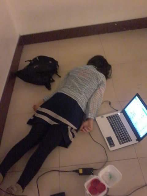 A young woman is lying flat on her face on the floor next to her laptop, looking defeated