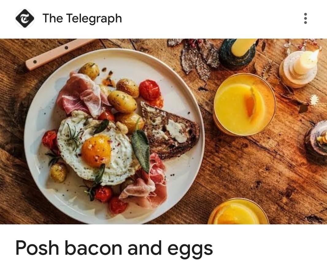 A top-down photograph shows a rustic breakfast laid out on a wooden table with shit (stuff) all over it, haphazardly. At the centre is a large white plate holding a “posh” version of bacon and eggs. On the plate is a sunny-side-up fried egg (probably ostrich or some nonsense) with a bright orange yolk, sprinkled with chilli flake. Around it are small roasted new potatoes with browned skins, roasted cherry tomatoes, and several thin, pink ribbons of prosciutto. A slice of dark, toasted sourdough bread with melted butter rests against the side of the plate. Sage leaves are scattered over the food.

To the right of the plate are two short glasses filled with orange juice, and slivers of what looks like orange peel.

At the top of the image is The Telegraph logo, and below the photo the headline reads: “Posh bacon and eggs.”
