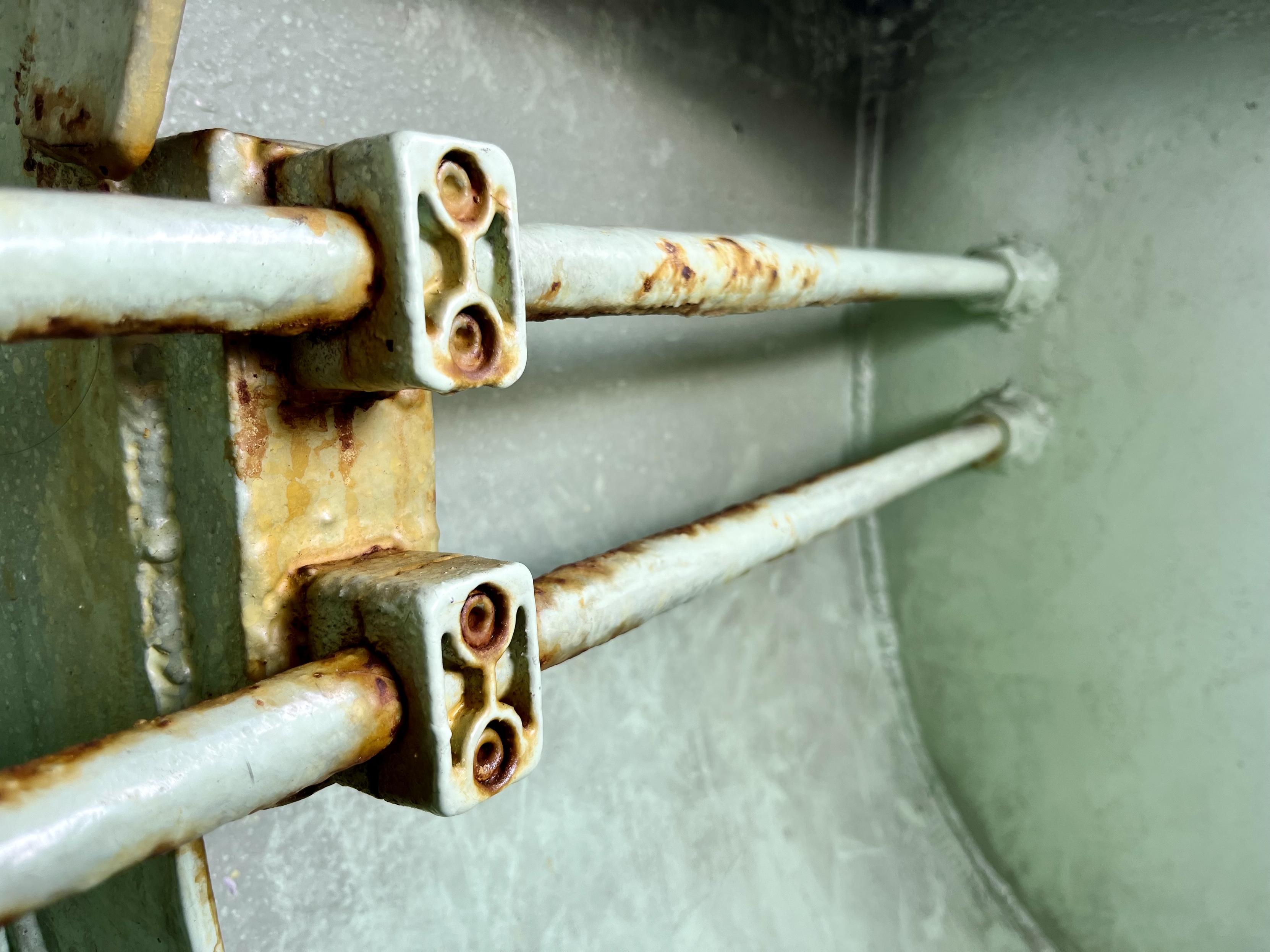 Detail photo of the metal inside wall of the prow of a vaporetto water bus. Two metal bars, held in place by two small blocks of metal which look like Lego pieces, run parallel in front of the gently curved metal side of the boat. Everything is painted a pale sage green - a very specific shade which is instantly recognisable as "vaporetto colour" - with patches of ochre-coloured rusty corrosion. It looks very steampunk retro-futuristic.