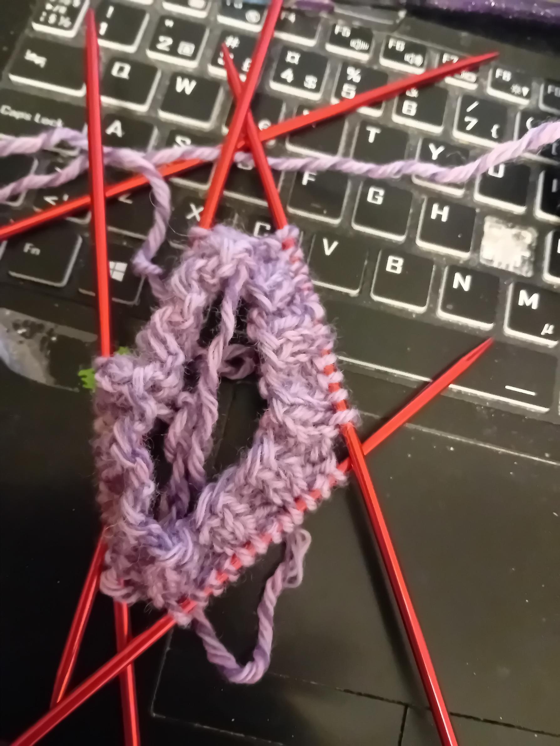 Pinkish-purple yarn on red DPN knitting needles, turning into the ribbing of a cuff