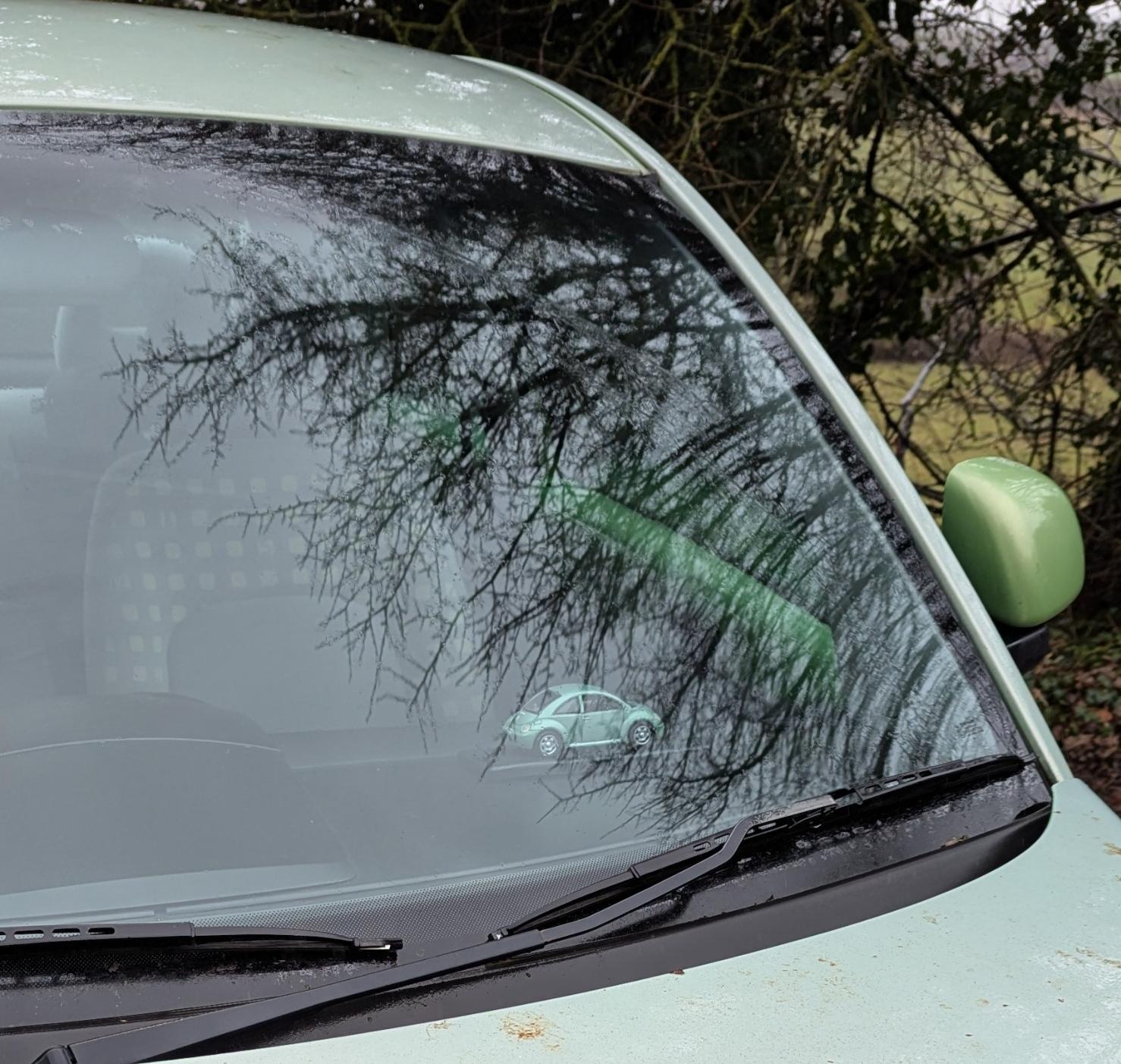 Photograph of a section of car windscreen, surrounded by pale green bodywork. A green metallic wing mirror is just visible. Inside the car, on the dashboard is a small replica Volkswagen Beetle toy car, viewed from the side.