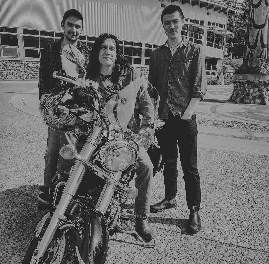 Three Indigenous men posing in front of a Songhees Wellness Center building and totem pole; the older man with long hair is seated on a motorcycle with two men standing on either side of him. Black and white image.
