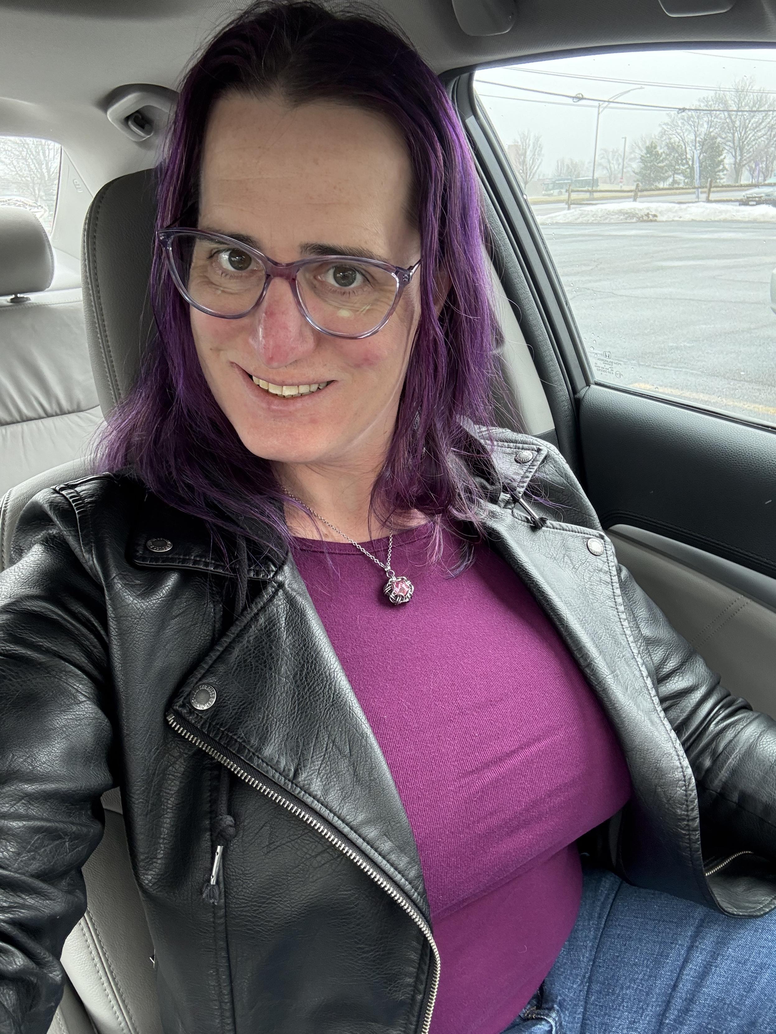 Selfie of a lady with purple hair, purple glasses, and purple blouse under a black leather jacket.