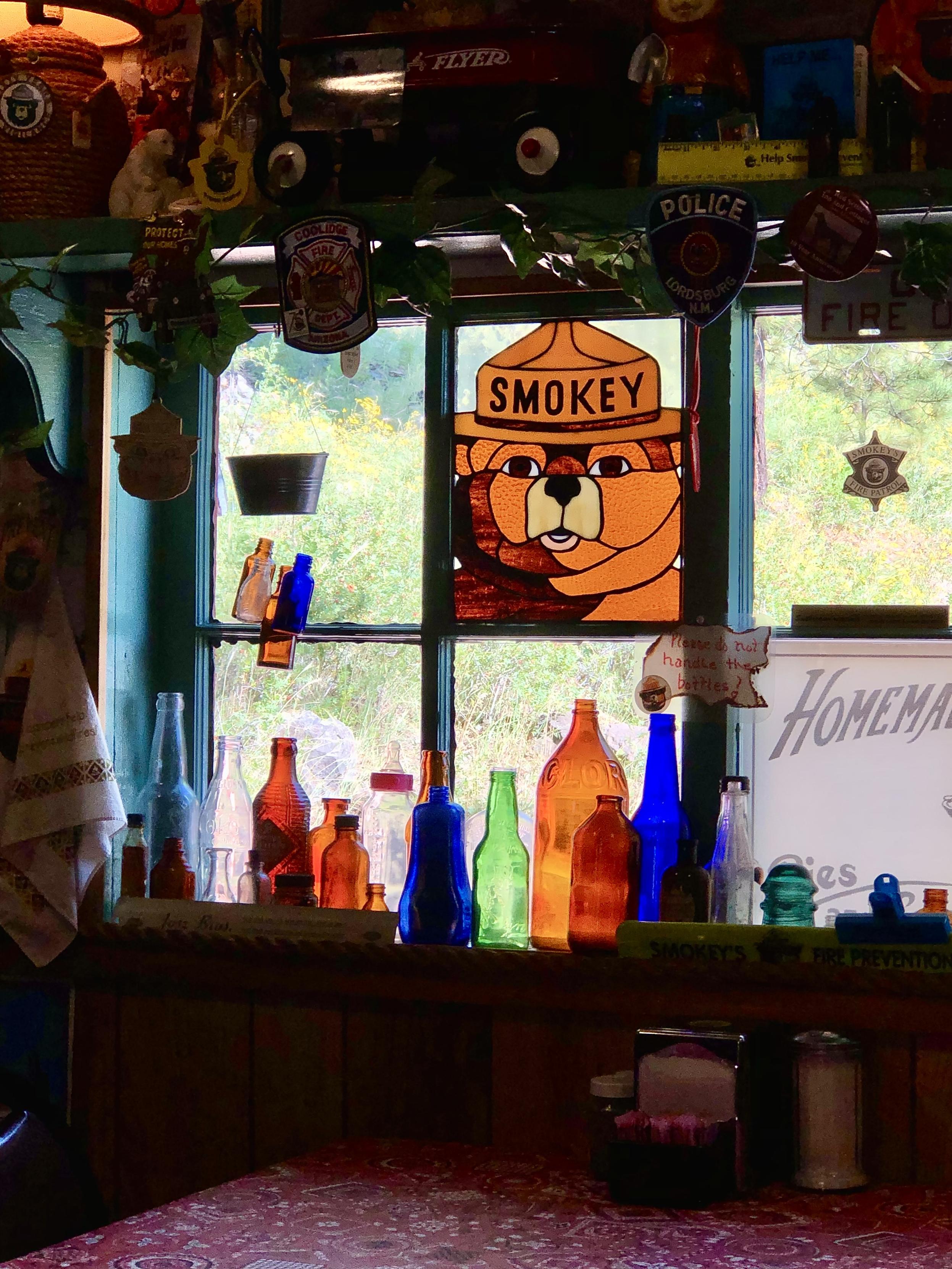 From inside looking out at the only cafe in rural Greer, Arizona. The light outside is bright and sunny so this table at the restaurant is inviting because there is a collection of antique glass bottles in various colors and the 6 panel window has something displayed in each. The center top has a stained glass brown bear head with the bear wearing a ranger hat that says SMOKEY on it. The inside is dark and cluttered but the outside is light. All the colored glass is glowing.