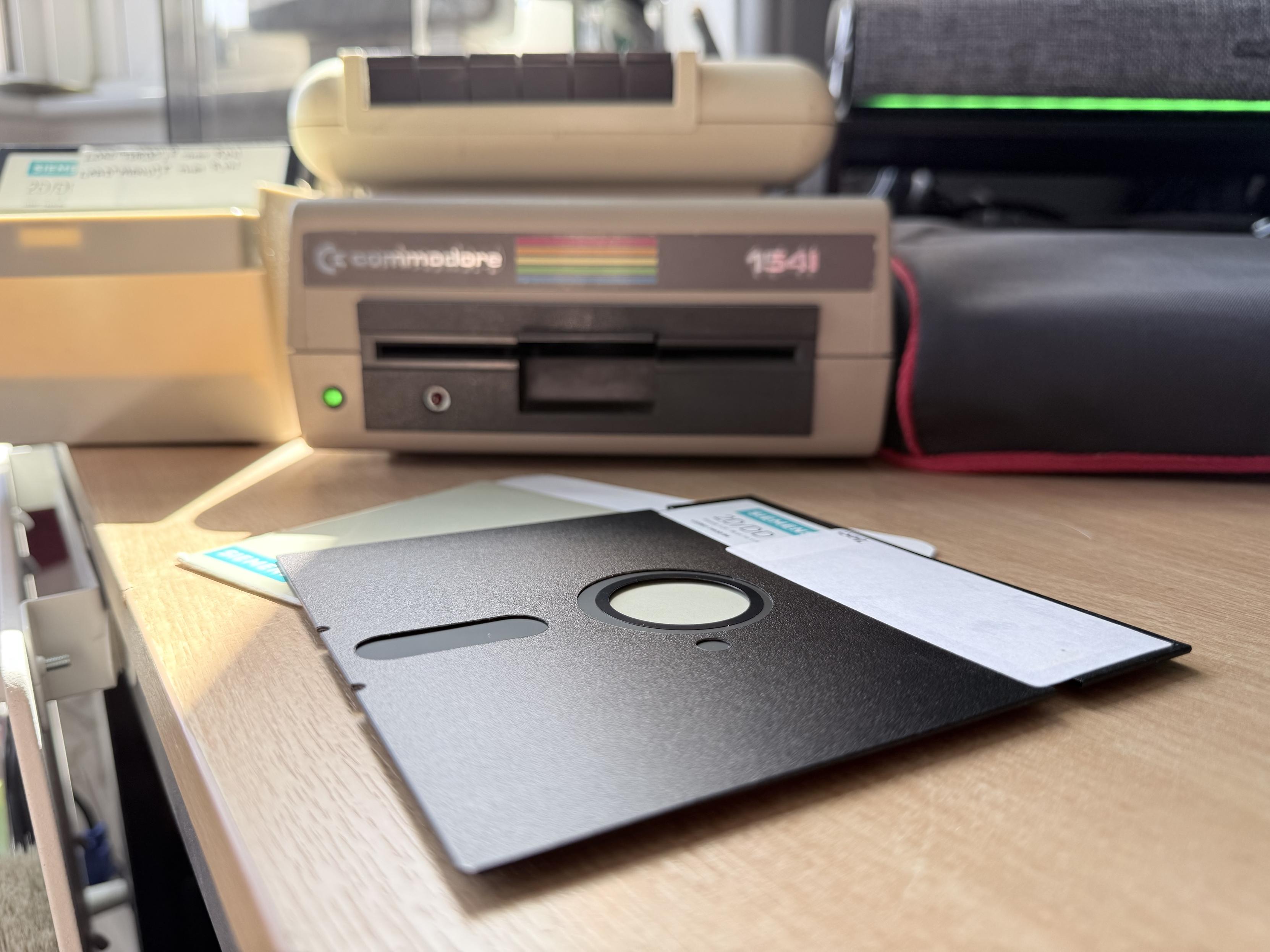 An image of a Commodore 1541 disk drive positioned on a wooden surface, with a black 5.25” floppy disk next to it. The drive features a green power indicator and is surrounded by a Commodore 64 Ultimate setup.