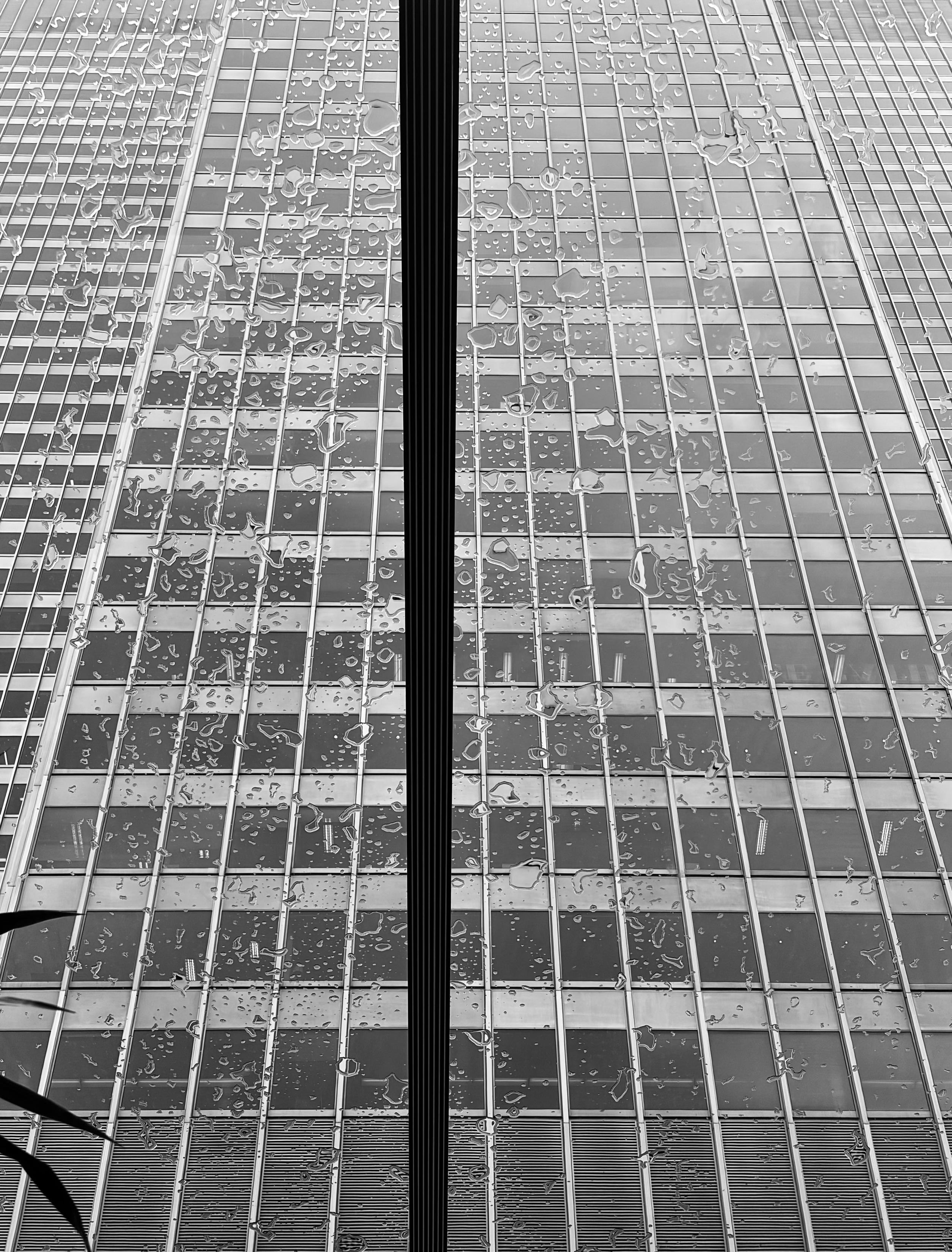 Black and white photograph of a tall concrete building with rows of windows, seen through a glass ceiling, covered with rain drops and splashes.