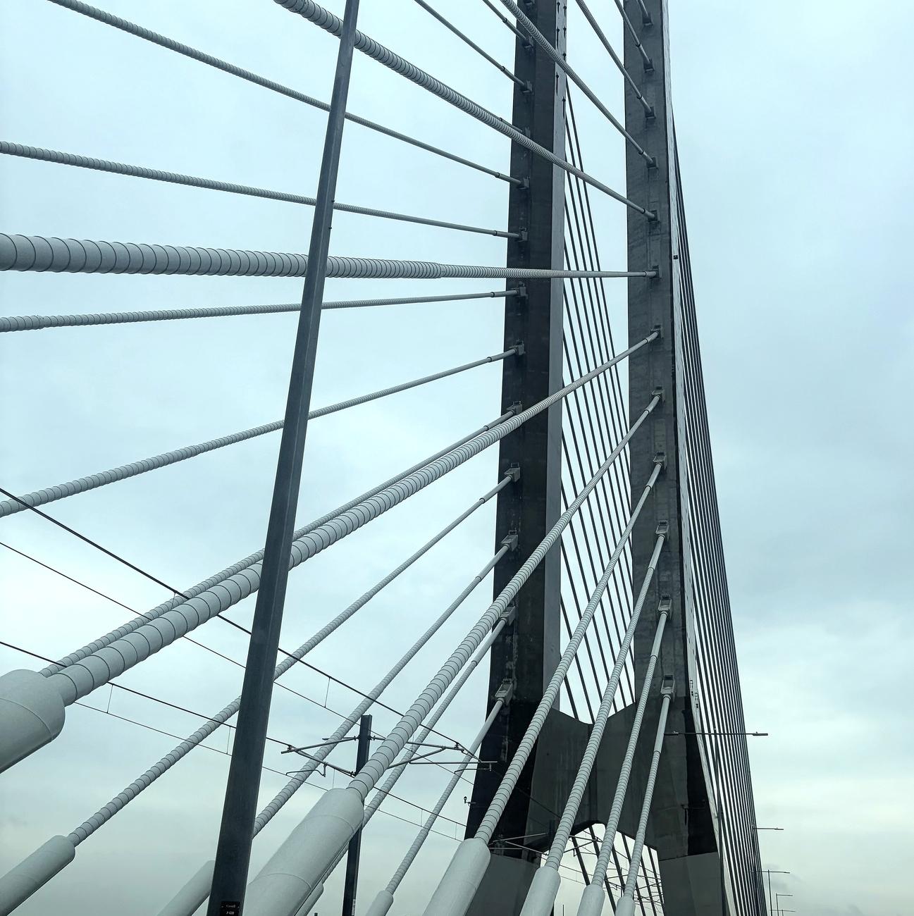 Photograph of a gray, two-part concrete bridge structure, forming a tall "U" shape, supporting white cables extending to the left of the image. Poles, lines, and lampposts are located below the structure and cables. The photo was taken while crossing the bridge, close to the right of the structure.

Photographie d'une structure grise en béton de pont en deux parties, formant une sorte de "U" très en hauteur, soutenant des câbles blancs s'étirant vers la gauche de la photo. Des poteaux et lignes ainsi que des lampadaires sont tout en bas sous la structure et les câbles. La photo a été prise en passant sur le pont, tout près à droite de la structure.