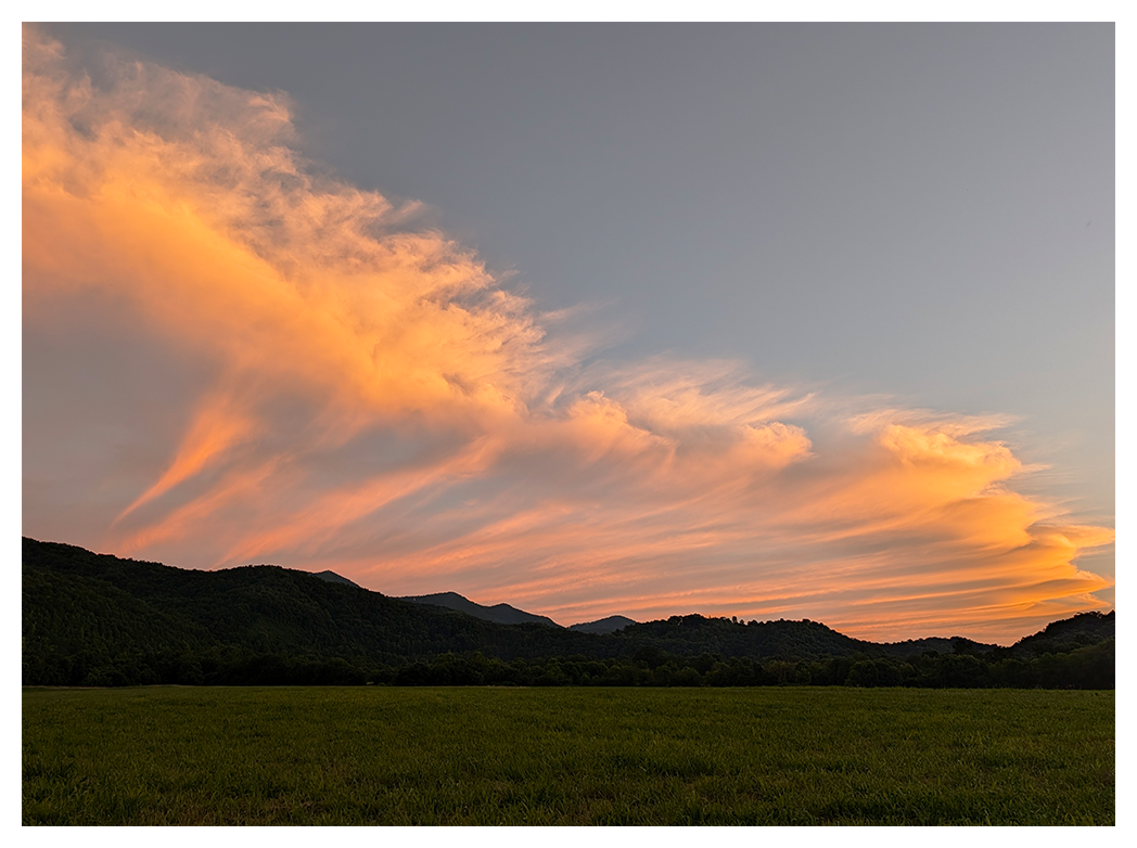 wispy orange clouds stretch across a pale blue sky at sunset above dark, tree-covered hills and a wide, green grassy field in the foreground.
