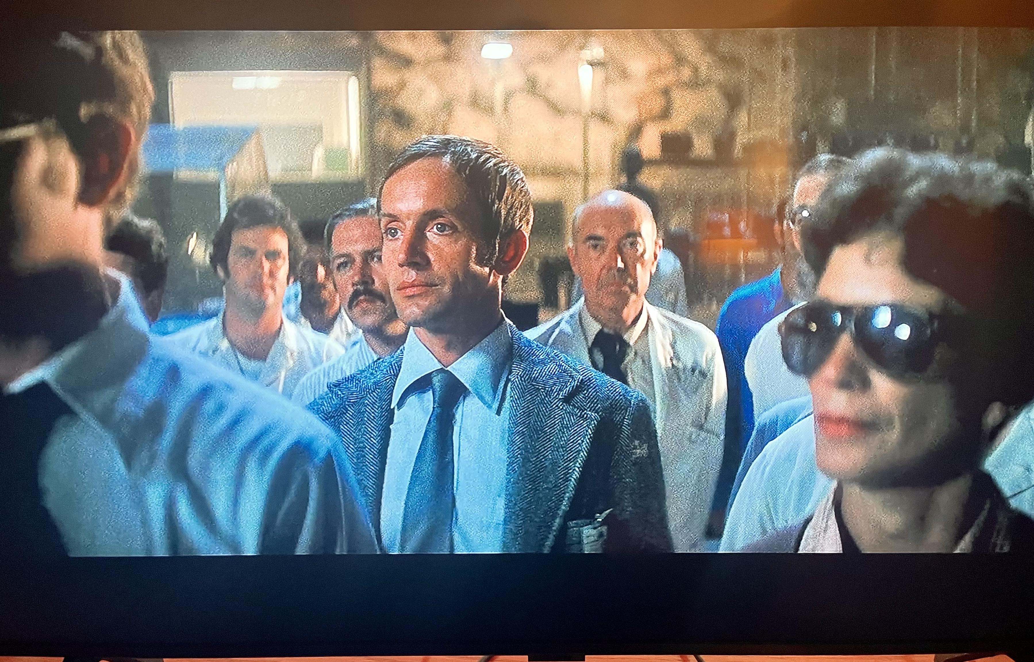 A group of people stands in a dimly lit setting. They appear focused and serious, with a variety of clothing styles, including lab coats and formal wear. The foreground features Lance in a blue suit