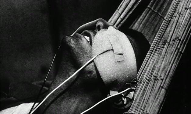 man in hammock with a mask on his face covering his eyes. the mask has wires attached. he seems to be in pain, mouth open slightly and head tilted back. a still from the film la jetée by chris marker.