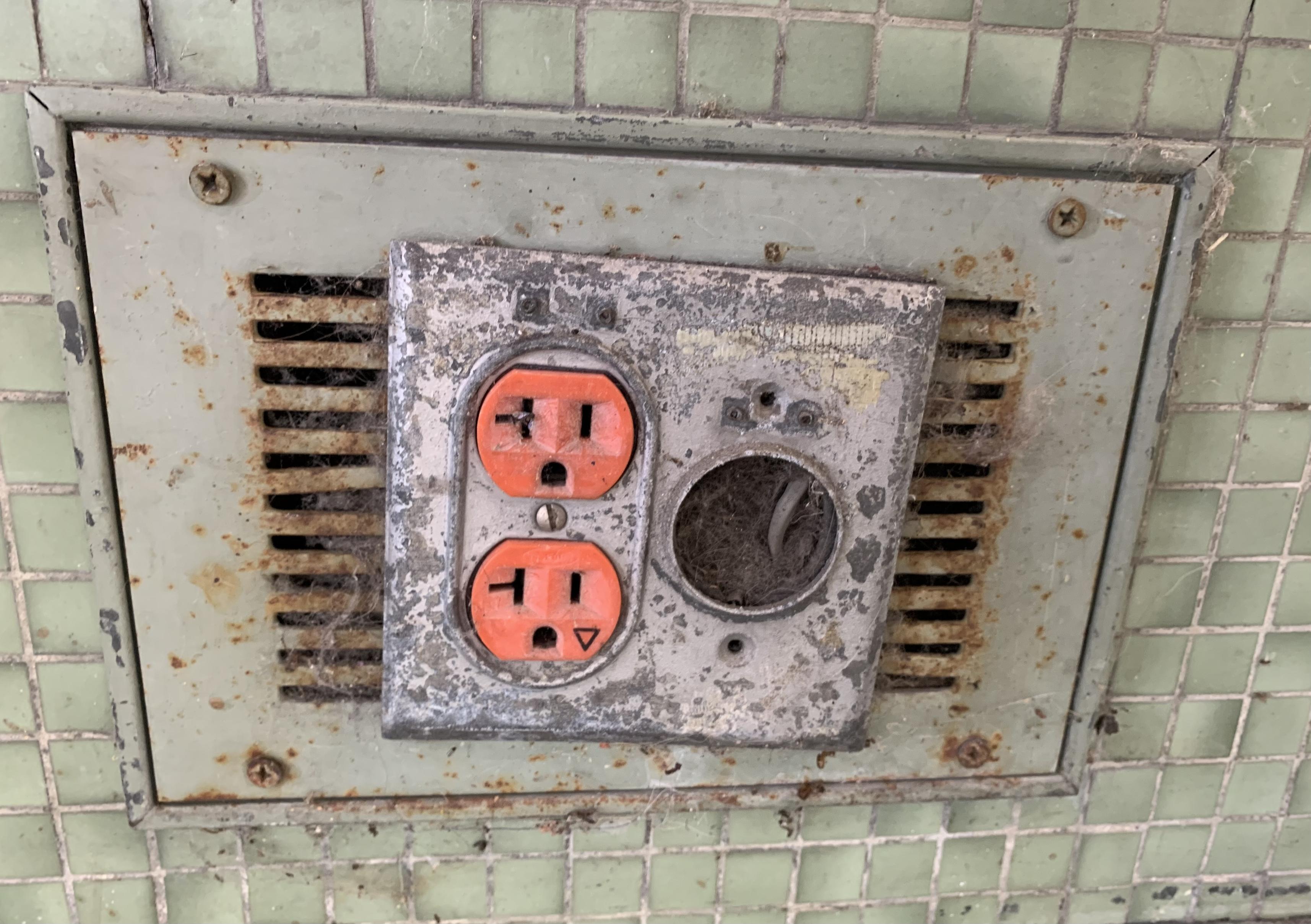 corroded power outlet with metal plate attached to rusty metal grate in green tiled wall — what is this?