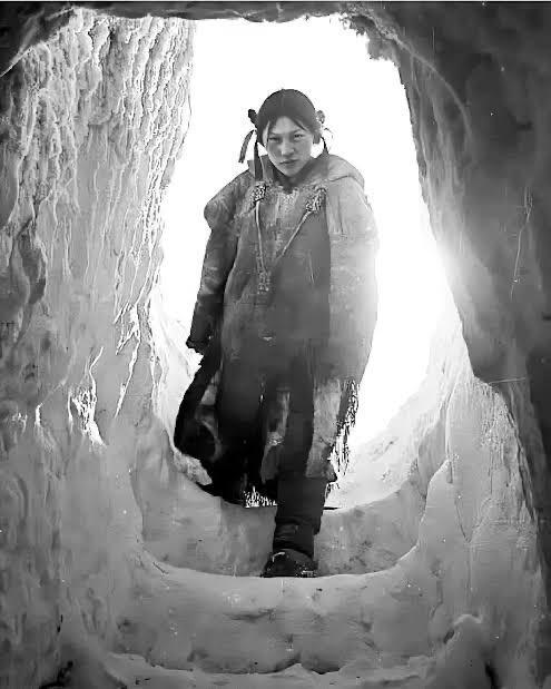 A black and white photo from the inside of an igloo, showing a teenaged Inuit girl entering.  The light comes from behind her — Helen Konek, photographed by Richard Harrington in 1949.
