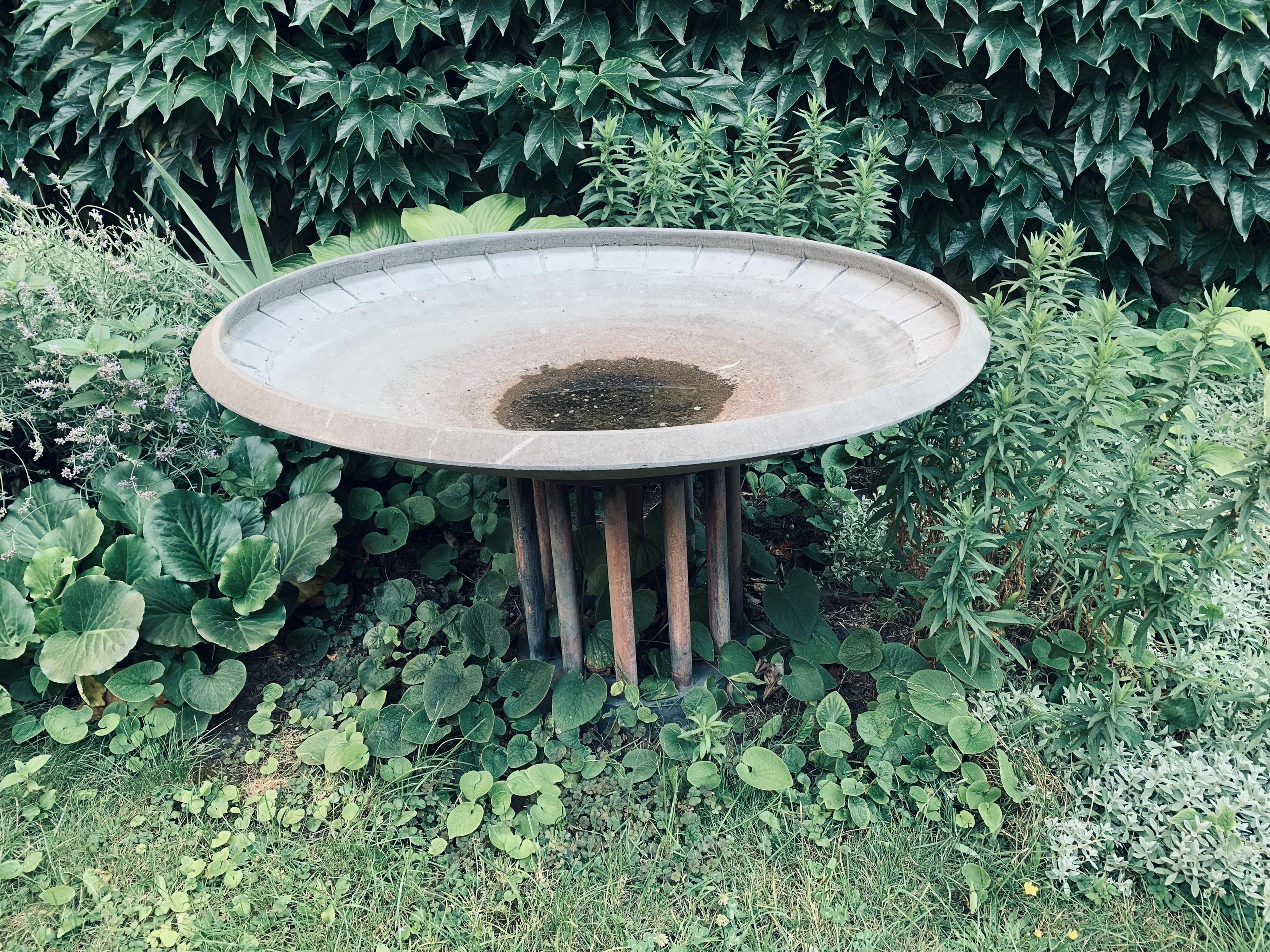 wide bird bath with a small pool of water at the base of the basin. it stands upon a circle of stout poles and is surrounded by greenery.