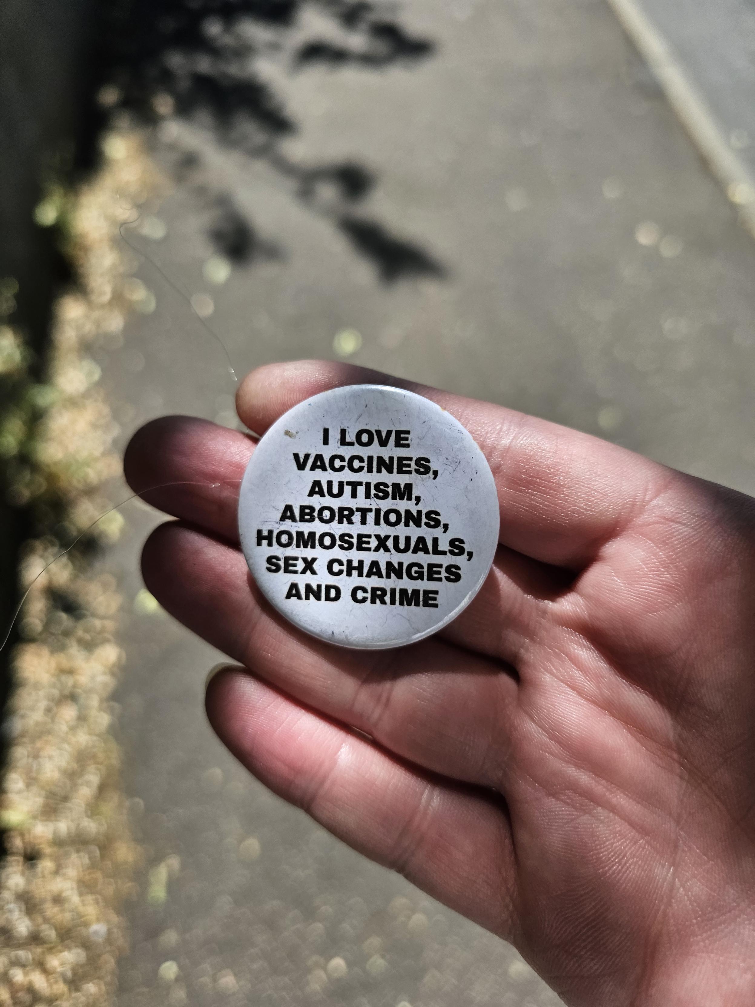 A photo of a white circular badge in my extremely white person hand. The badge says, in block capitals, I love vaccines, autism, abortions, homosexuals, sex changes, and crime.