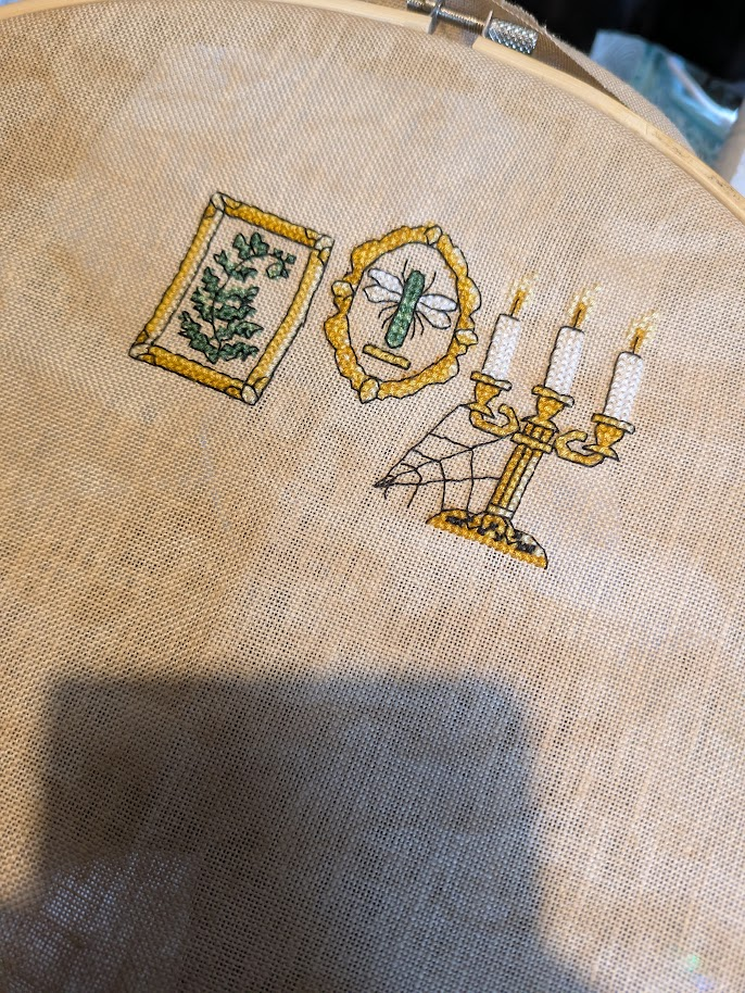 Cross stitch photo. Not So Scary Apothecary. A golden picture frame with a green fern in it. A dragonfly is in an oval golden frame. Then a golden candelabra with three white candles and a cobweb.