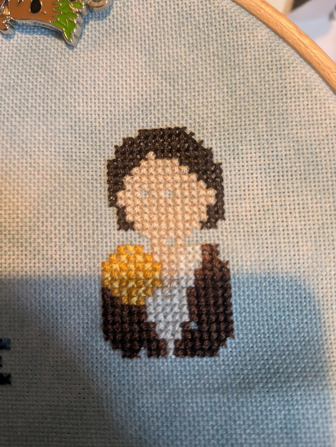 Cross stitch of a person with dark hair, wearing a brown jacket and white top, a golden ball is at their shoulder. The pattern is not complete, the person does not have any eyes or a mouth yet, looking rather eerie.