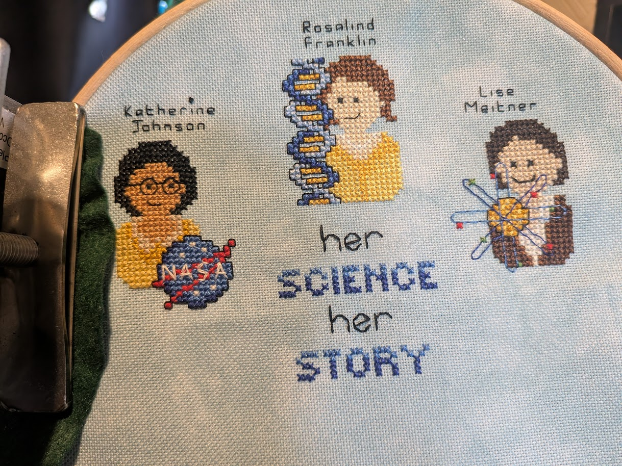 Cross stitch of Her Science, Her Story.  From left to right we have Katherine Johnson with the NASA logo, Rosalind Franklin with a DNA helix and now Lise Meitner with a very simplified atom.