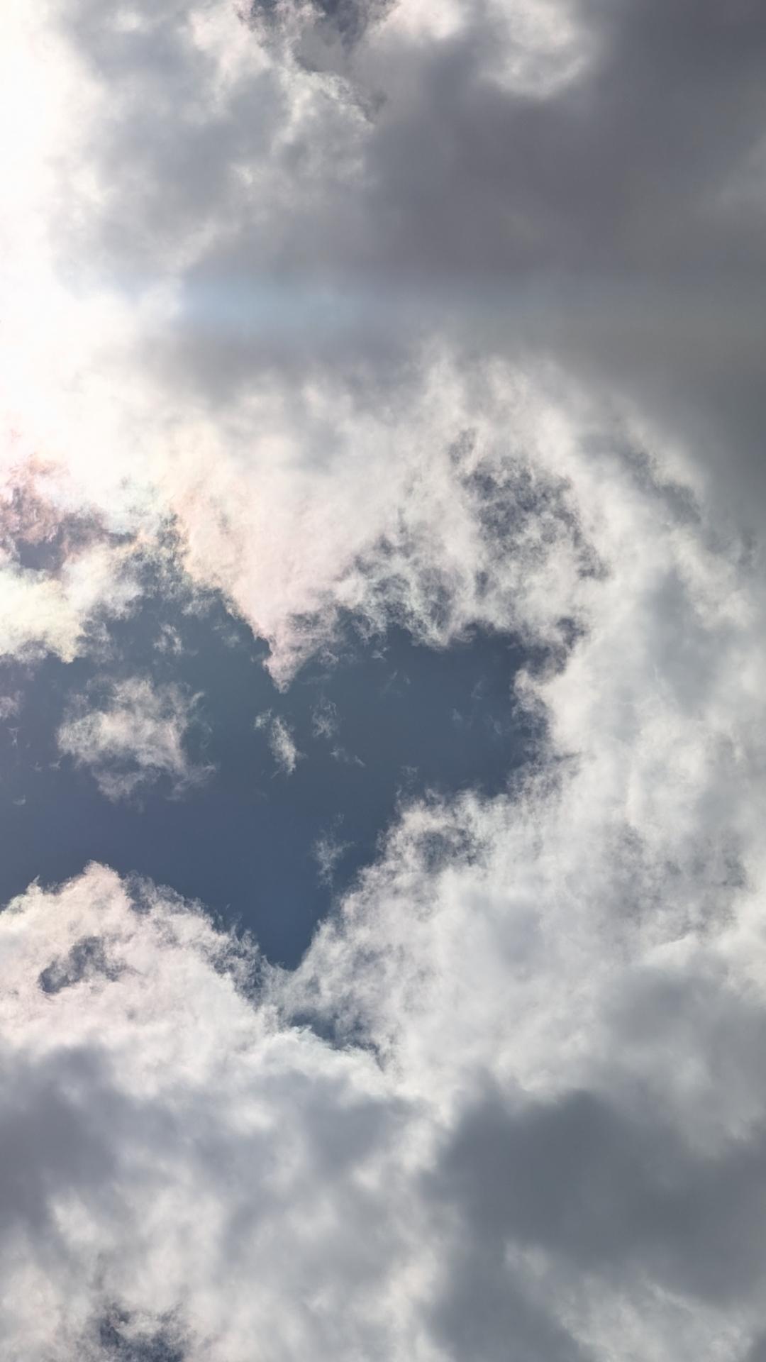 A photo of the clouds in the sky, with deep grey-blue sky peeking through the edges of the whispy clouds, forming a heart shape. The clouds surrounding the heart shape have grey centers, with brilliant white edges. A lens flare shines across the top of the photo, from the sun in the top left, towards the right edge. Taken from West Jordan, Utah.