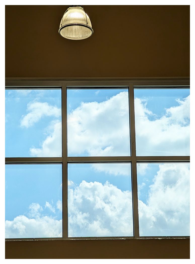 A ceiling light hangs above a large window divided into six panes. Bright blue sky and fluffy white clouds are visible through the glass, filling the window with daylight.