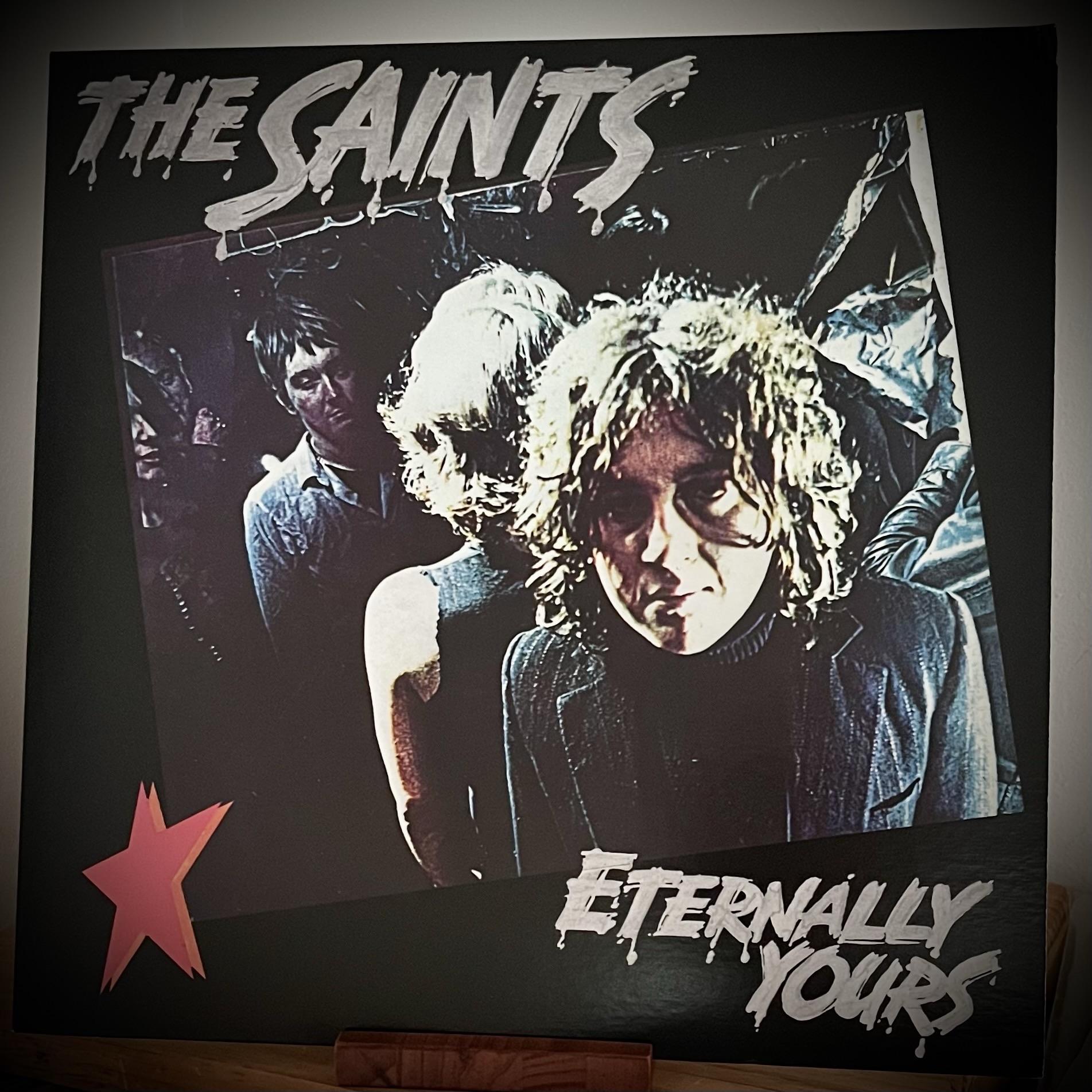 The image features the album cover of "Eternally Yours" by The Saints. It shows a moody, dark photograph of band members with one prominently displayed in the foreground. The title "THE SAINTS" is at the top, and the title is at the bottom.
