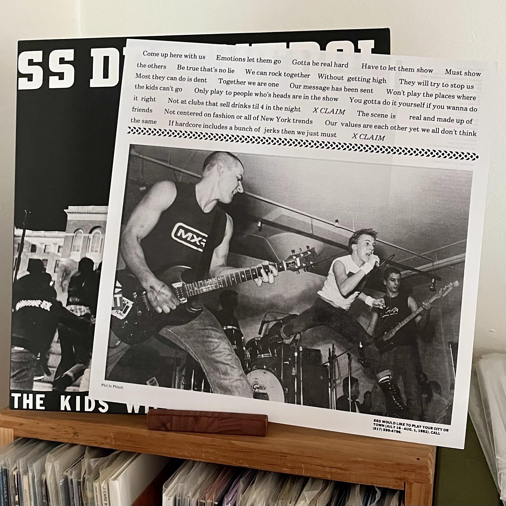 The image features a close-up of SS Decontrol Kids Will Have There Say displayed on a wooden shelf, with a black-and-white photograph of a band performing. The band consists of a guitarist and a vocalist, both in dynamic poses, with another musician visible in the background.