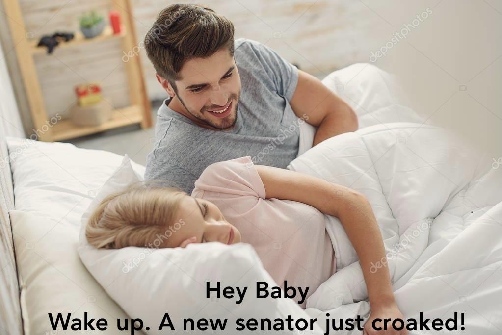 A young man is playfully waking up a young woman in bed, both relaxed and cozy under a blanket. Text reads: “Hey Baby wake up a new senator just croaked.”