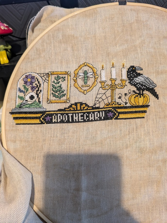 Cross stitch photo. The top shelf of the Not So Scary Apothecary is complete. The shelf is black and gold with the word apothecary in white in the centre of it. From left to right are a bell jar with birds skull in it, a golden rectangular frame with a green fern, a small oval frame with a dragonfly in it, a candelabra with three tall white candles, a spiders web goes from the candles to the shelf, finally a large black and grey bird stands on a pumpkin.