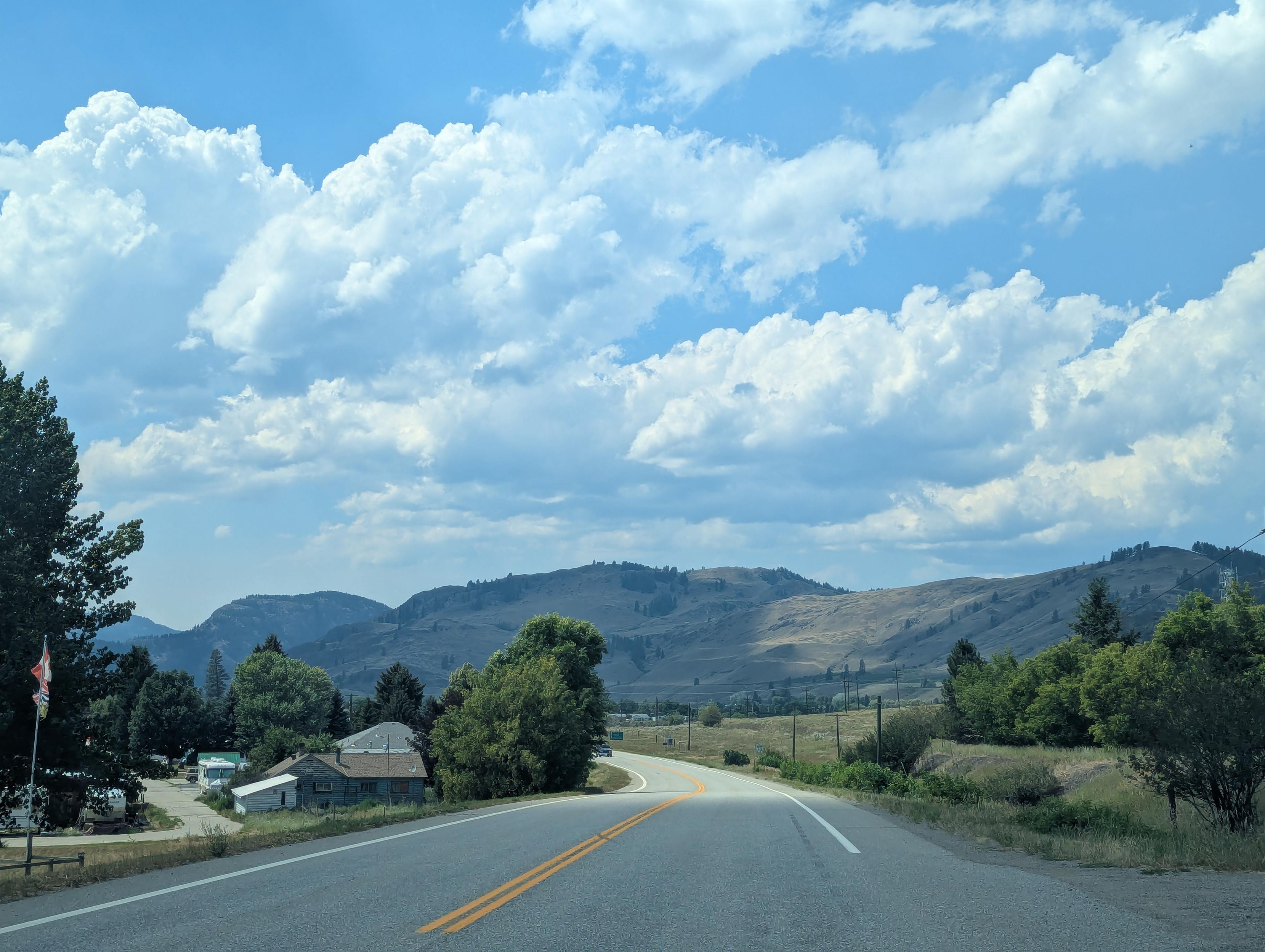 Big fluffy white clouds against some arid mountains. We're in a car driving on a winding highway sparsely lined with trees.