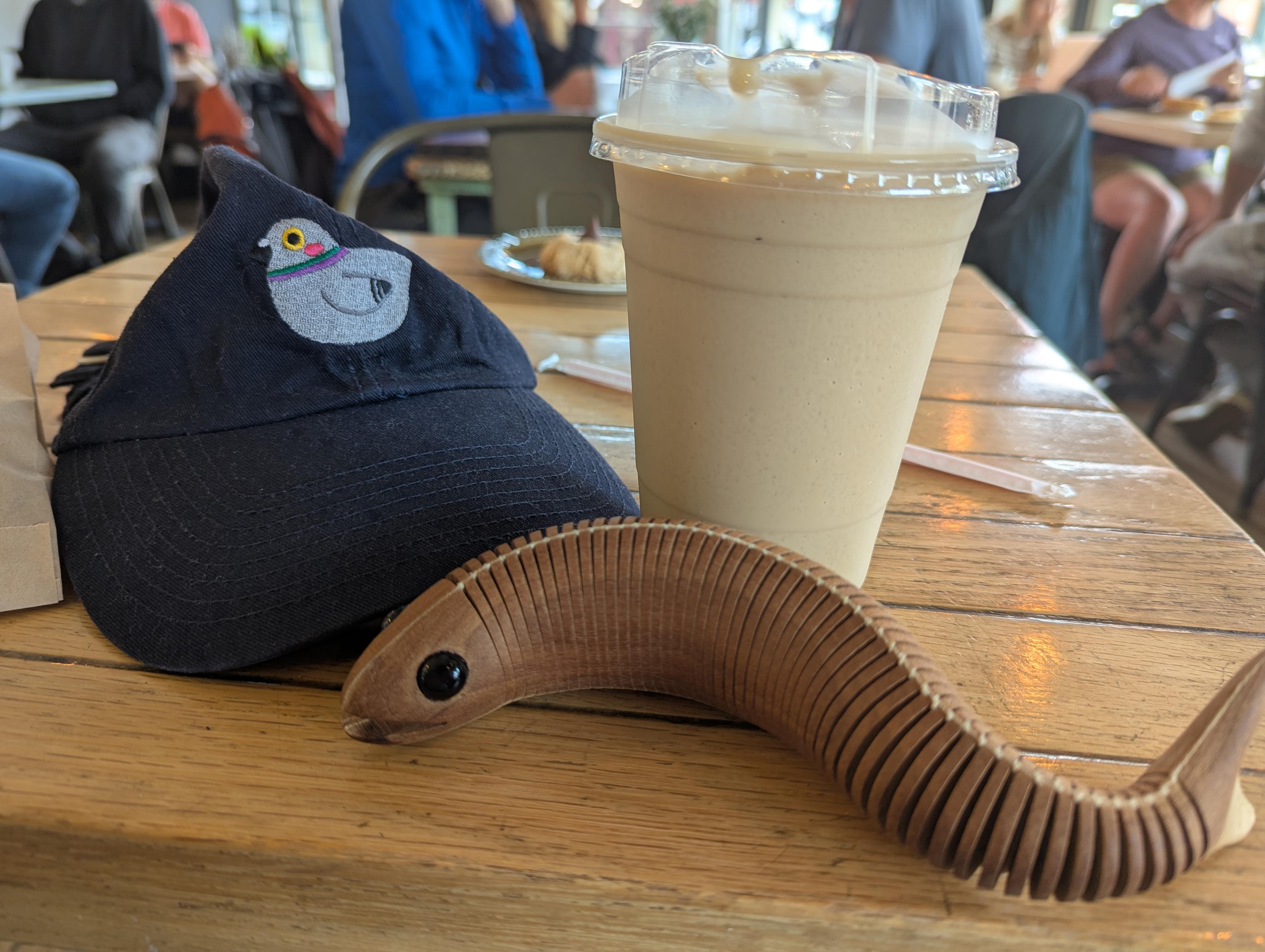 My travel kit: my baseball cap featuring a silly embroidered pigeon, an iced coffee beverage, and Pitupaq, my traveling wooden fidget fish from Cape Breton.
The photo is taken in a busy coffee shop. No faces are visible.