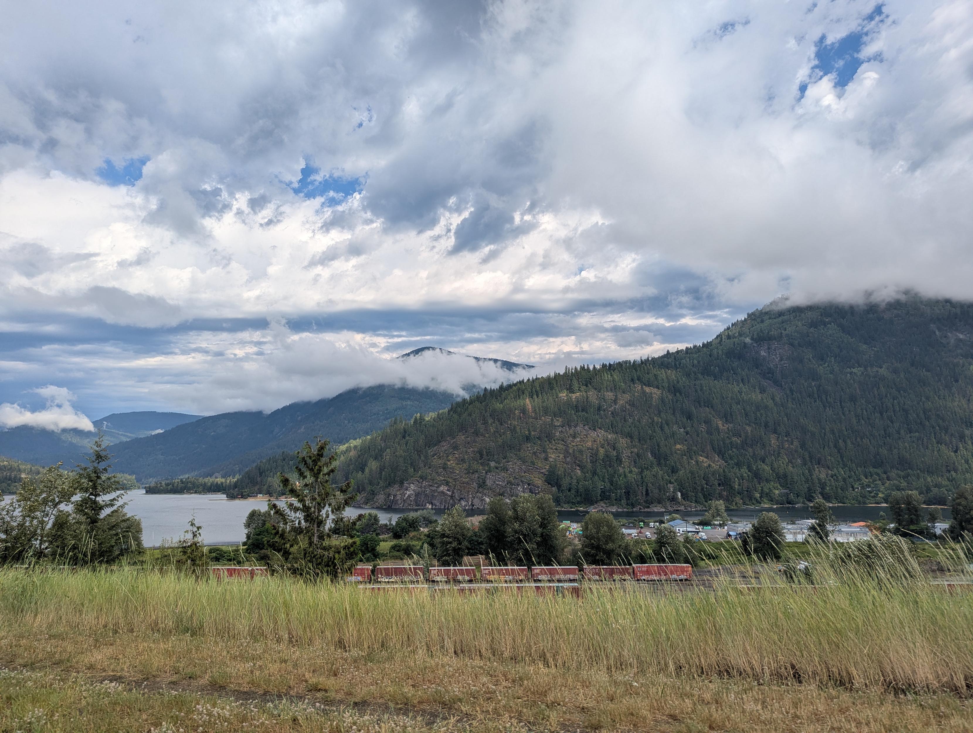 Landscape photo showing layers of typography. From closest to the photographer: hay fields, a train track, a row of trees, a lake, low clouds, a low mountain, fluffy clouds, another mountain, another mountain,