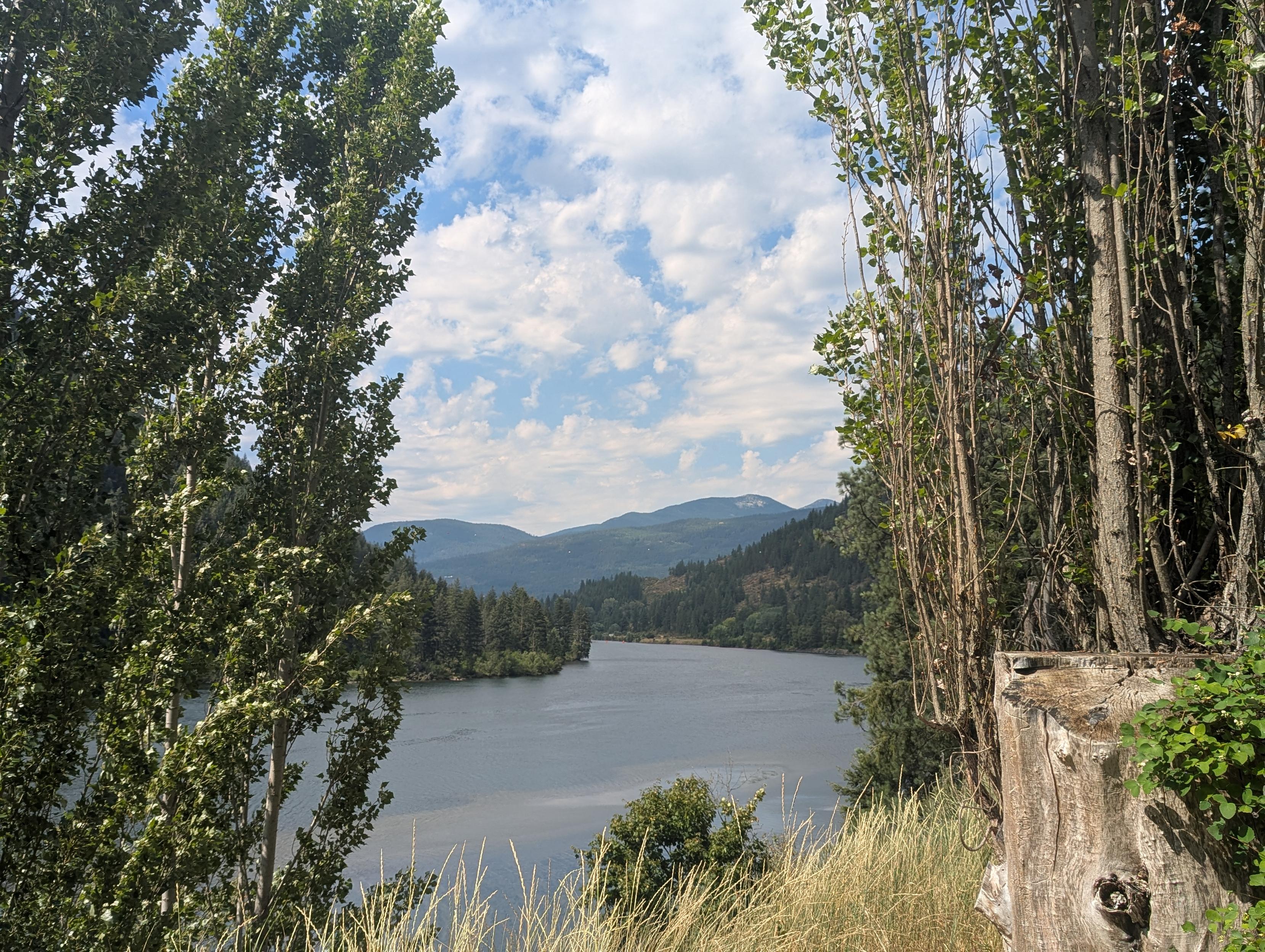 A lake in the mountains bordered by trees and a large stump in the foreground. The blue sky peeks out from behind the large fluffy white clouds.