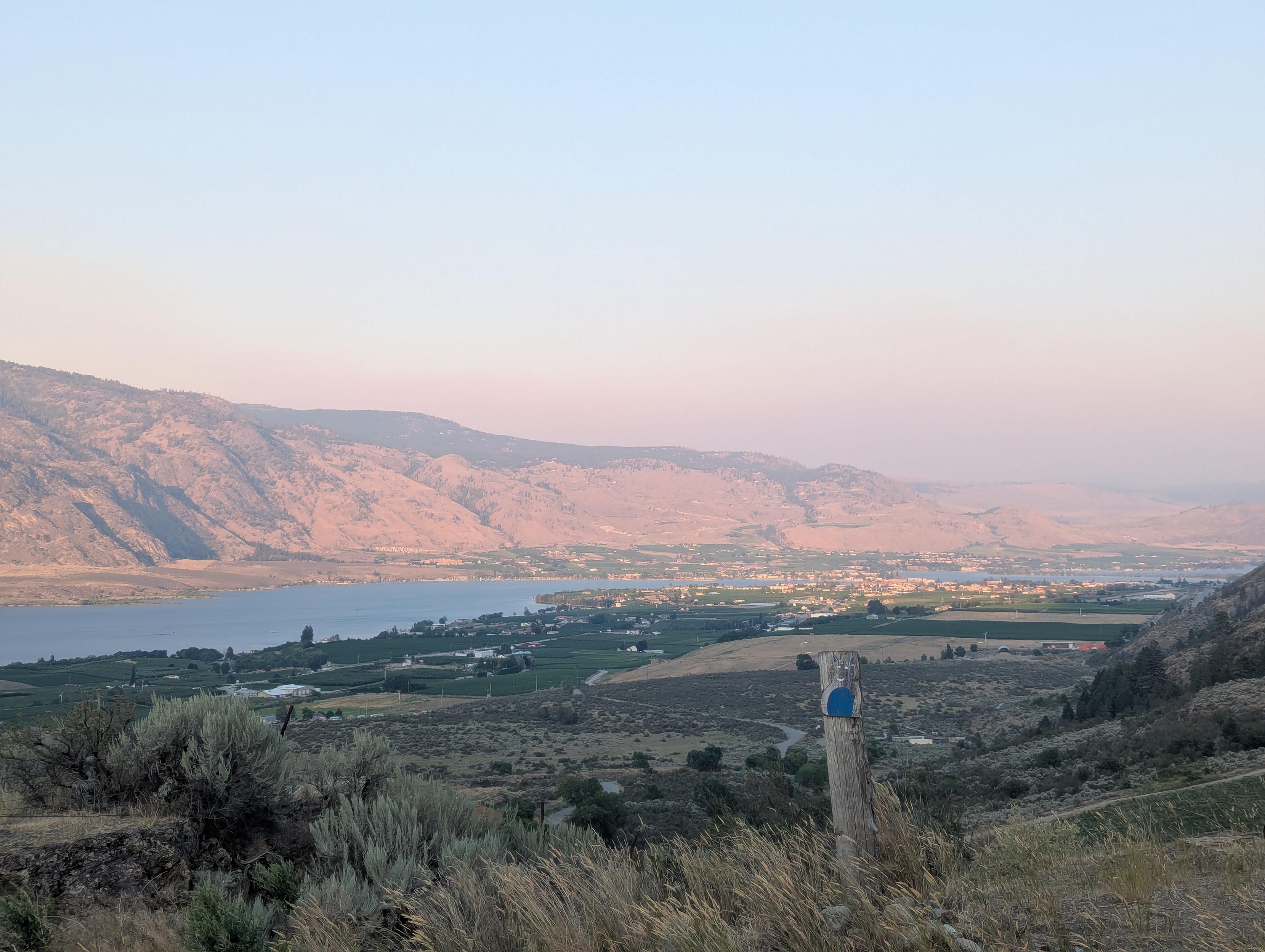 A long desert mountain sits beyond a long lake through the smokey orange haze. A small city can be seen in the distance, with some wineries on this side of the river. There's a post in the foreground.