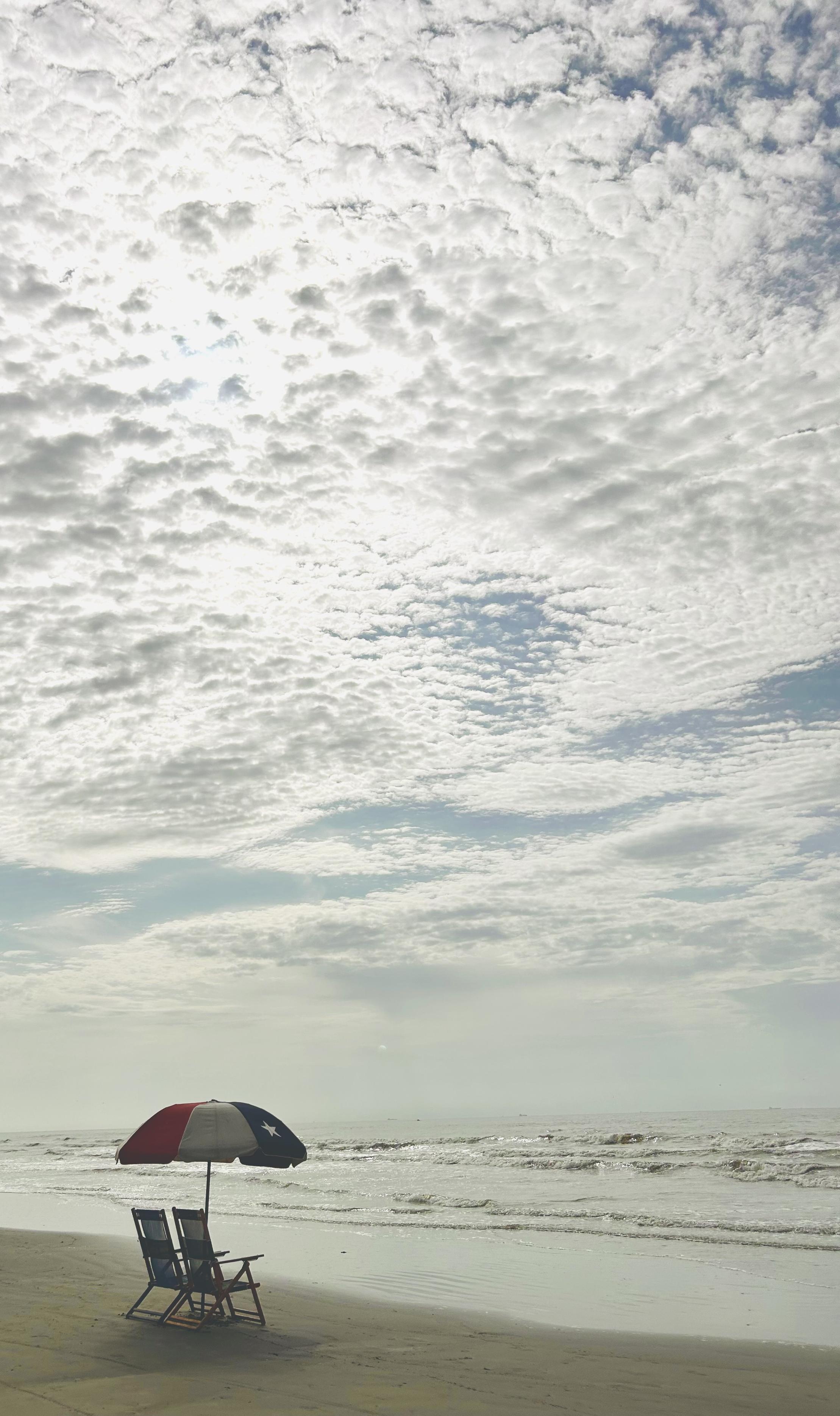 Two chairs under one umbrella that is facing the ocean. A cloudy sky