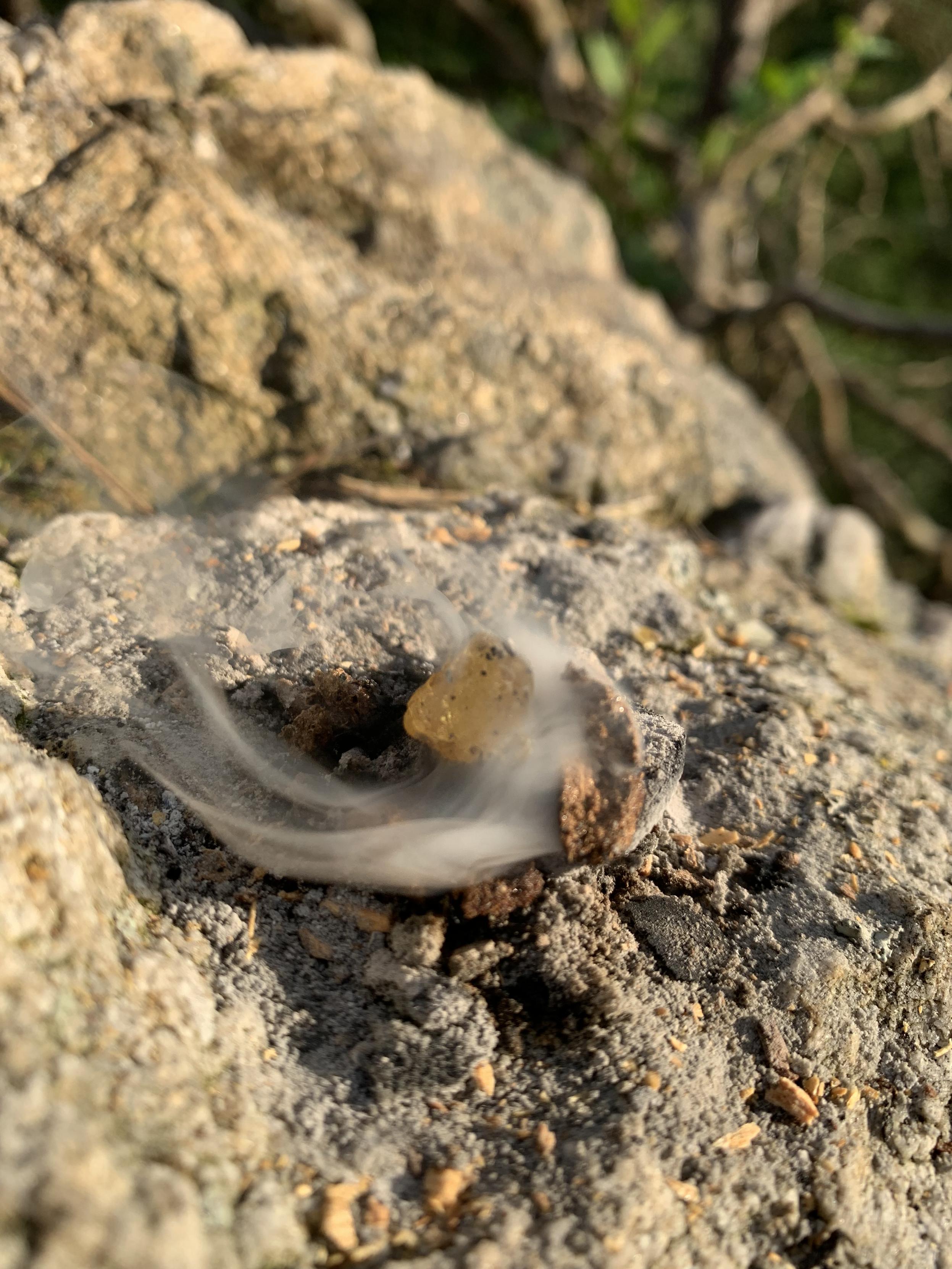 a piece of amber being consumed by a swirl of smoke upon a rock