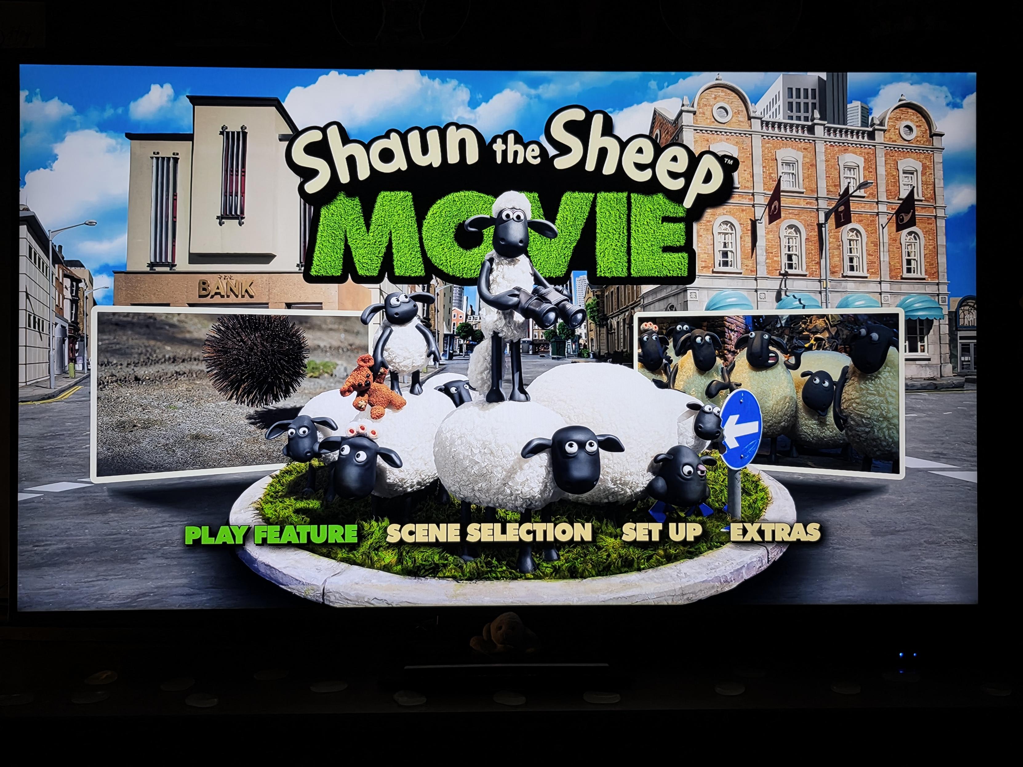 A photo of a television screen displaying the main menu for the Blu-ray of *Shaun the Sheep Movie*.
In the center of the menu, set against a bright city street background with brick buildings under a sunny blue sky, the iconic flock is adorably piled onto a small, circular grassy roundabout. Shaun stands triumphantly at the very top of the sheep stack, looking key-focused through a pair of binoculars. Little Timmy sits perched on another sheep's back, happily clinging to his tiny orange teddy bear, while the rest of the flock huddles underneath with their delightfully wide, innocent eyes.
Above the flock sits the movie title logo: "Shaun the Sheep" in classic white cartoon lettering, with "MOVIE" prominently displayed beneath it in giant, grass-textured green block letters. Flanking the central sheep pile are two rectangular preview boxes showing clips from the film—the left displaying a funny spiky hairpiece/urchin on the street, and the right showing the wide-eyed flock gathered together in awe.
Along the bottom curve of the grassy patch, the interactive menu choices are listed in bold lettering: "PLAY FEATURE" (glowing in bright green, ready for a cozy movie night), "SCENE SELECTION", "SET UP", and "EXTRAS".
