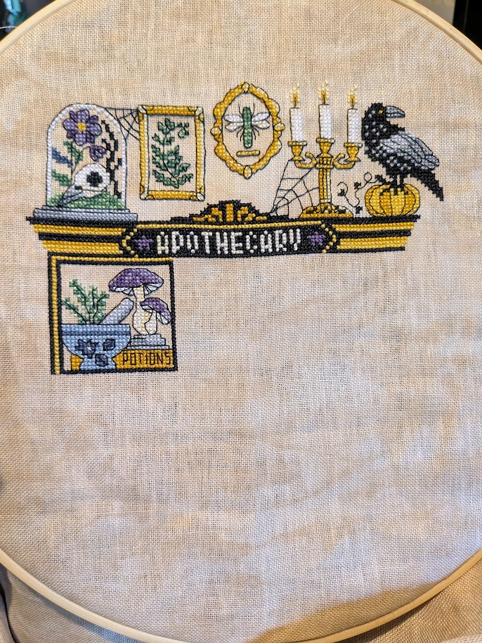 Cross stitch photo. We are onto the inside of the cabinet now. Along the top of the apothecary cabinet are a bell jar, fern, dragonfly, candleabra, and a Raven on a pumpkin. Below the bell jar is a cabinet square, inside this section is a silvery blue pestle and mortar with some green plant in it, next to that are two purple and white mushrooms, there is also a golden label with the word potions on it.