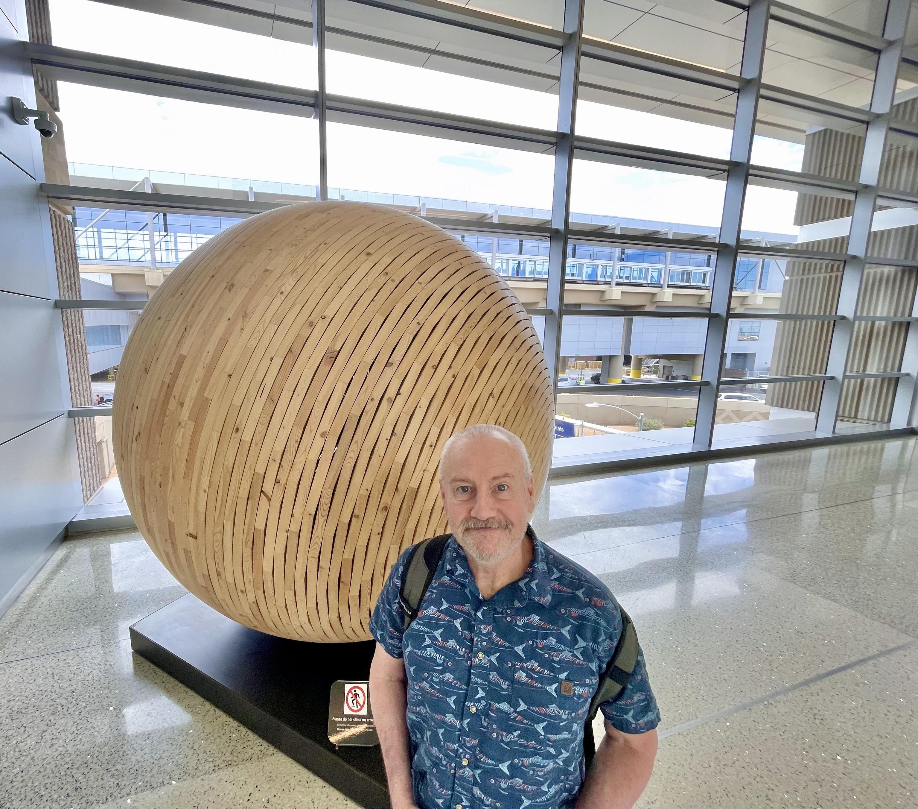 A man stands in front of a large ball made of curved strips of wood. In the background is a large window behind a rectangular metal grid.